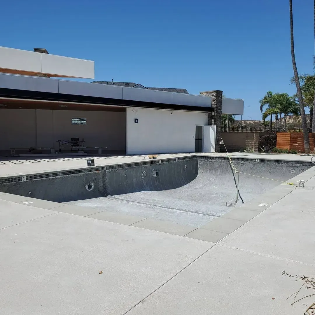 Empty, in-progress swimming pool in front of a modern building under a clear, blue sky.