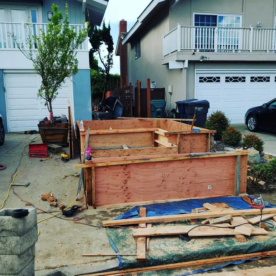 Construction site in a driveway. Wooden forms are built, a blue tarp and tools are scattered. Houses are in the background.