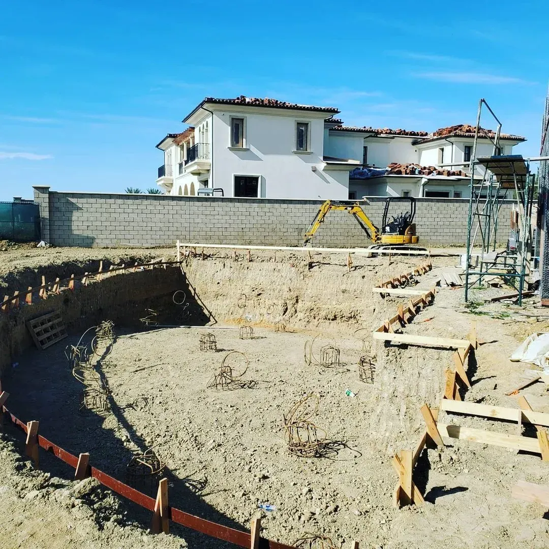 Pool excavation in progress at a residential construction site. A house and blue sky are in the background.