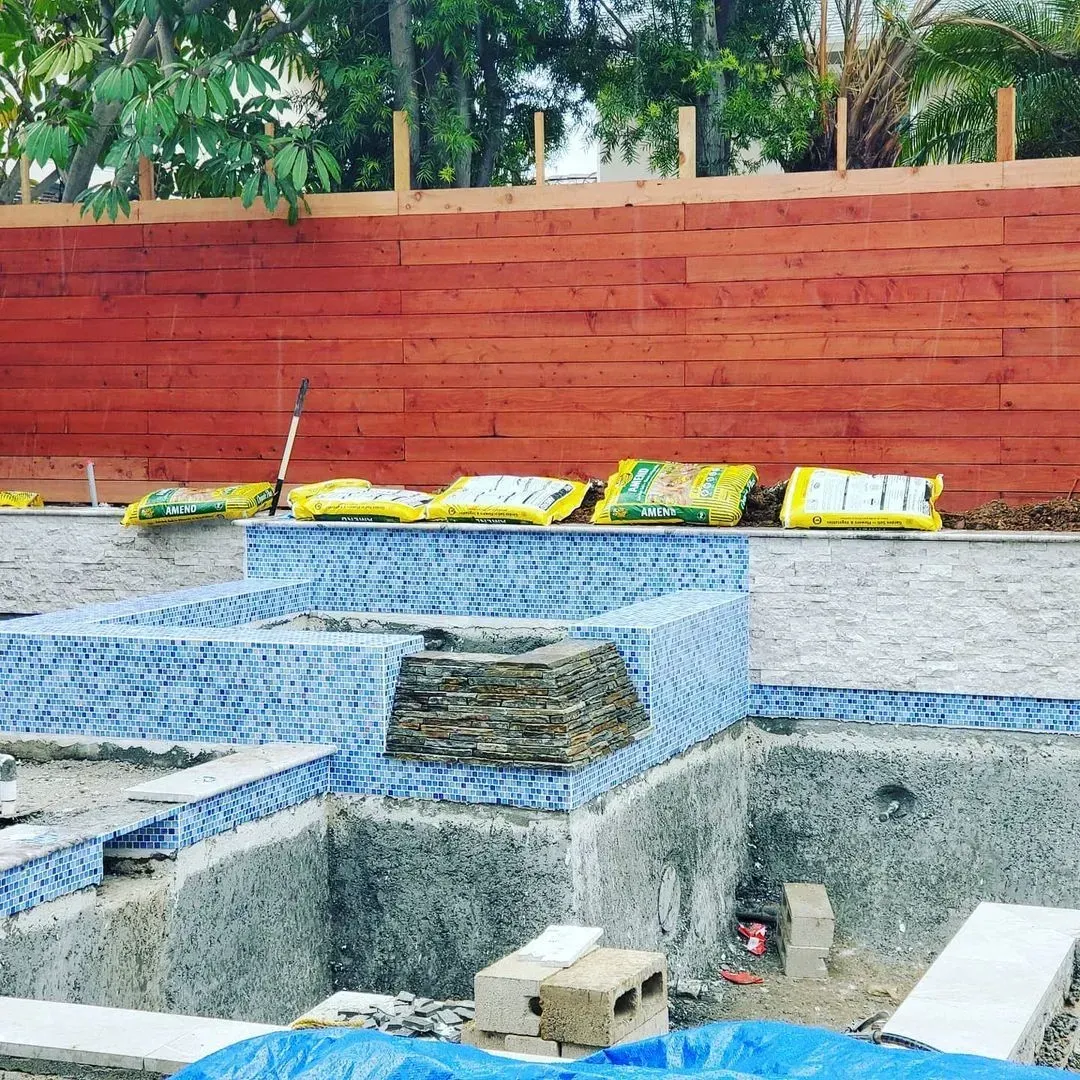 Pool under construction with blue tile, red wooden fence, and bags of material.