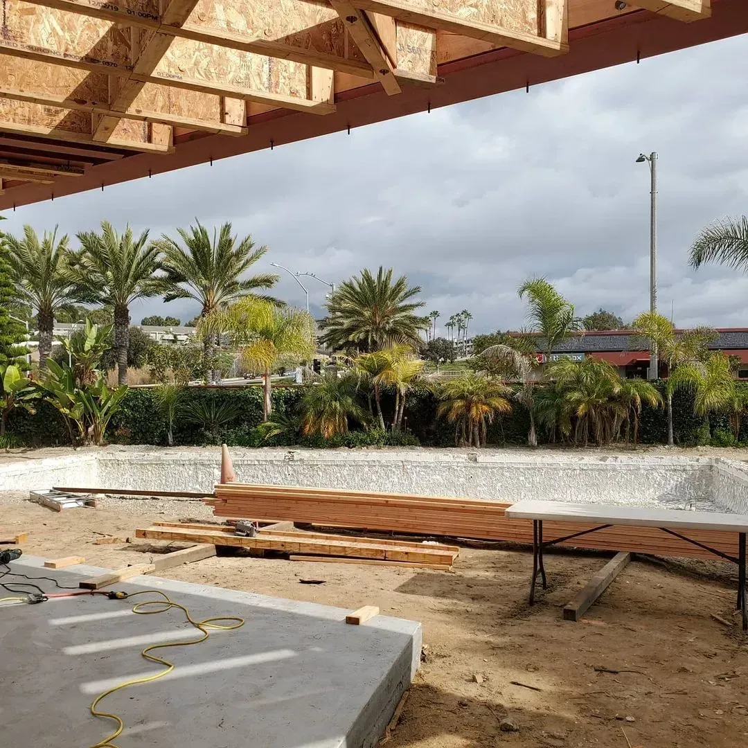 Construction site with unfinished pool, wooden beams, palm trees, and cloudy sky.