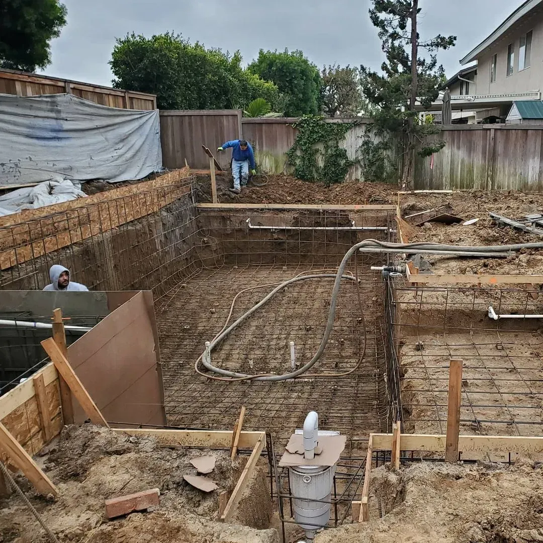 Pool construction site: partially excavated rectangular shape, workers, wood framing, dirt, cloudy sky.
