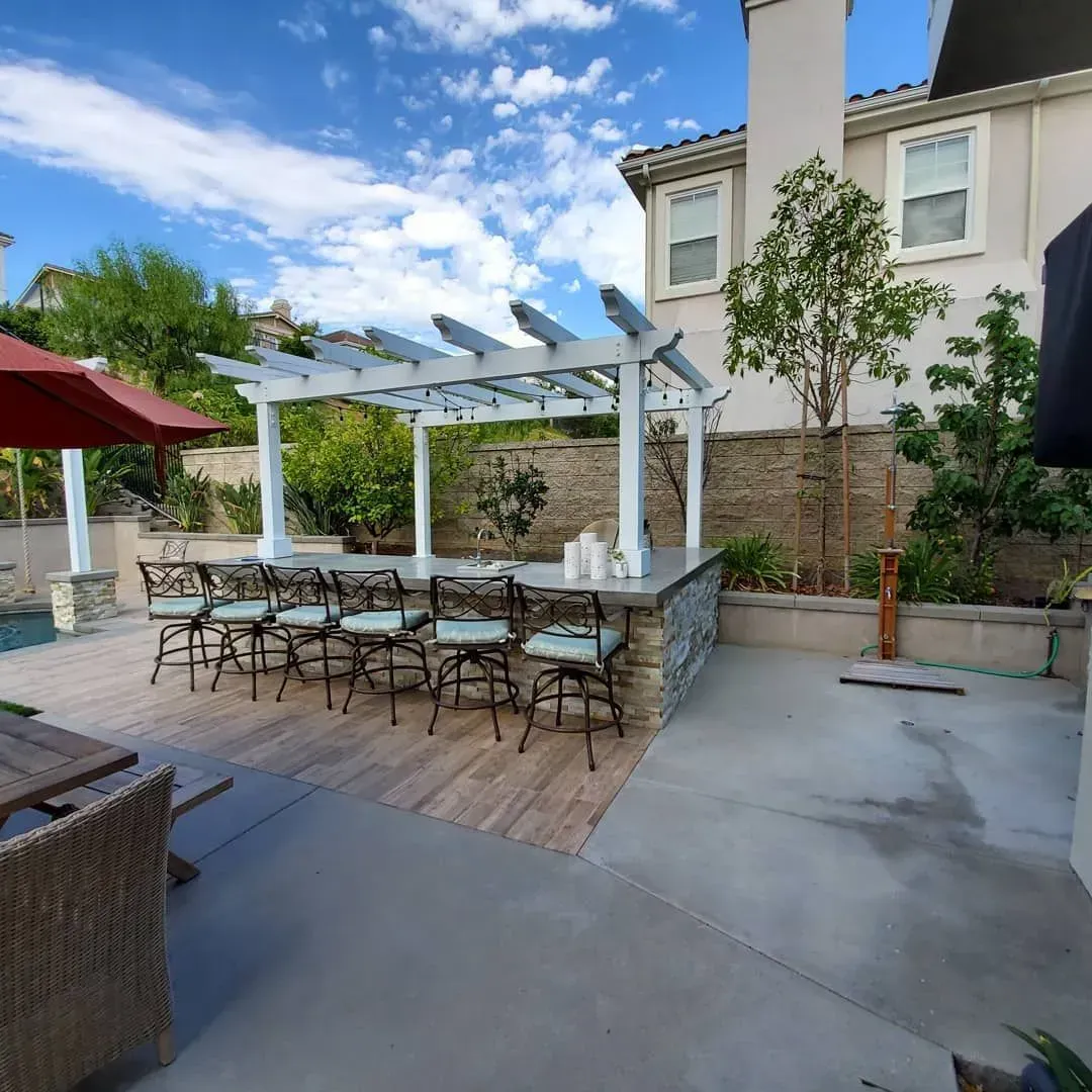 Outdoor bar with seating under a pergola, beside a pool and house, on a sunny day.