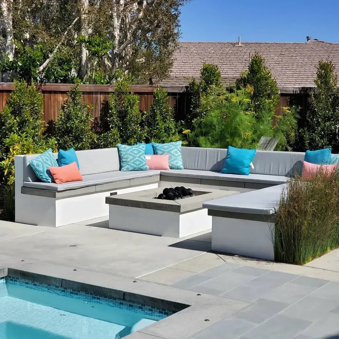 Outdoor seating area with fire pit and pool, surrounded by greenery; blue, gray, and coral accents.