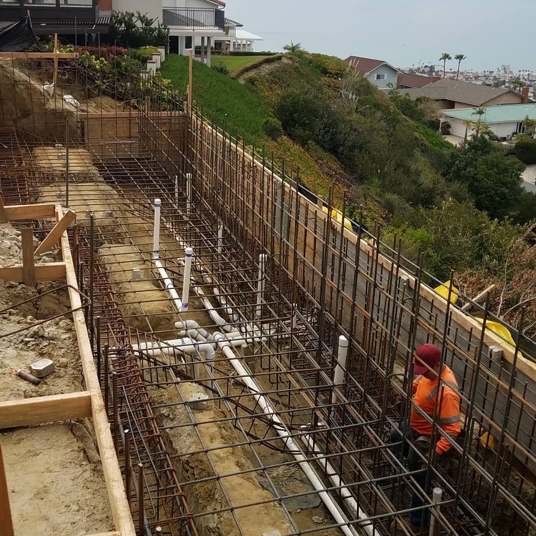 Construction site with a worker in orange vest, rebar framework, and plumbing. Houses on a hillside in the background.
