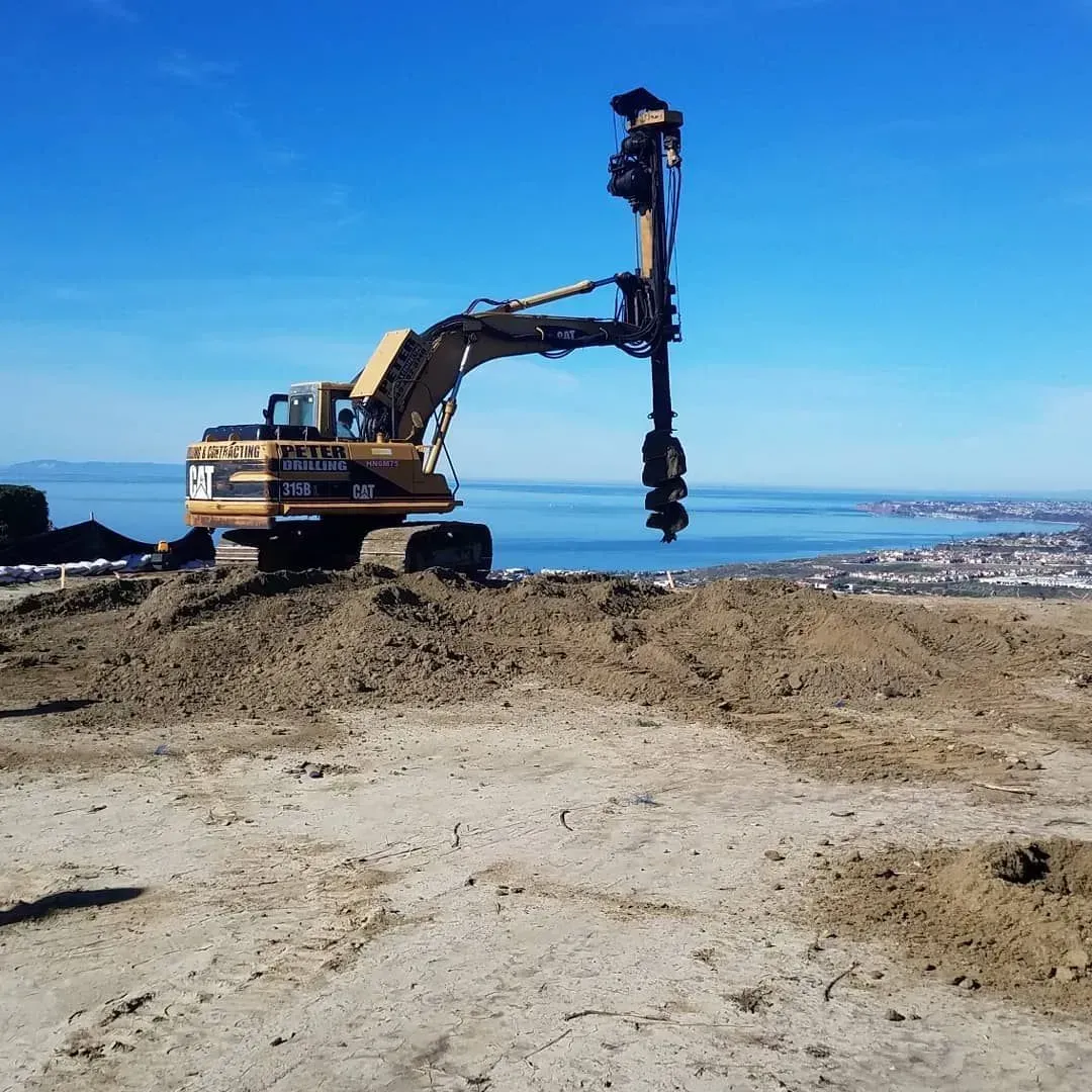 Excavator with drill attachment on a dirt mound, blue sky, water, and cityscape in the background.