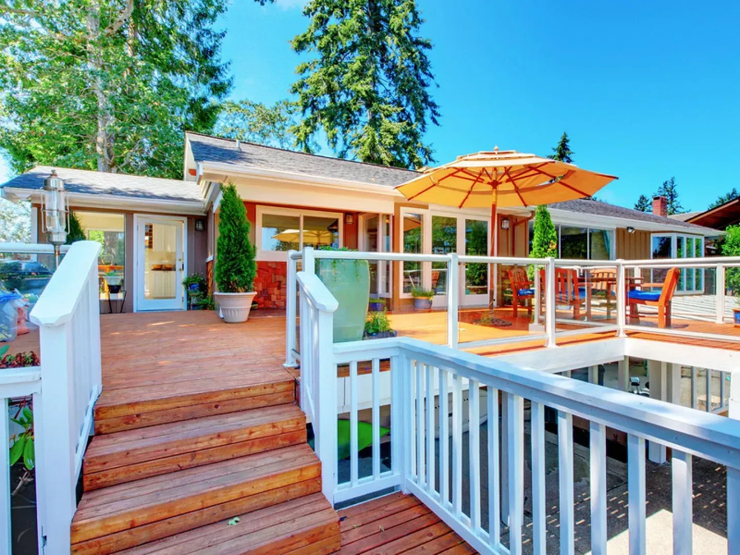 Wooden deck with stairs, white railings, and an open umbrella overlooking a house with windows.