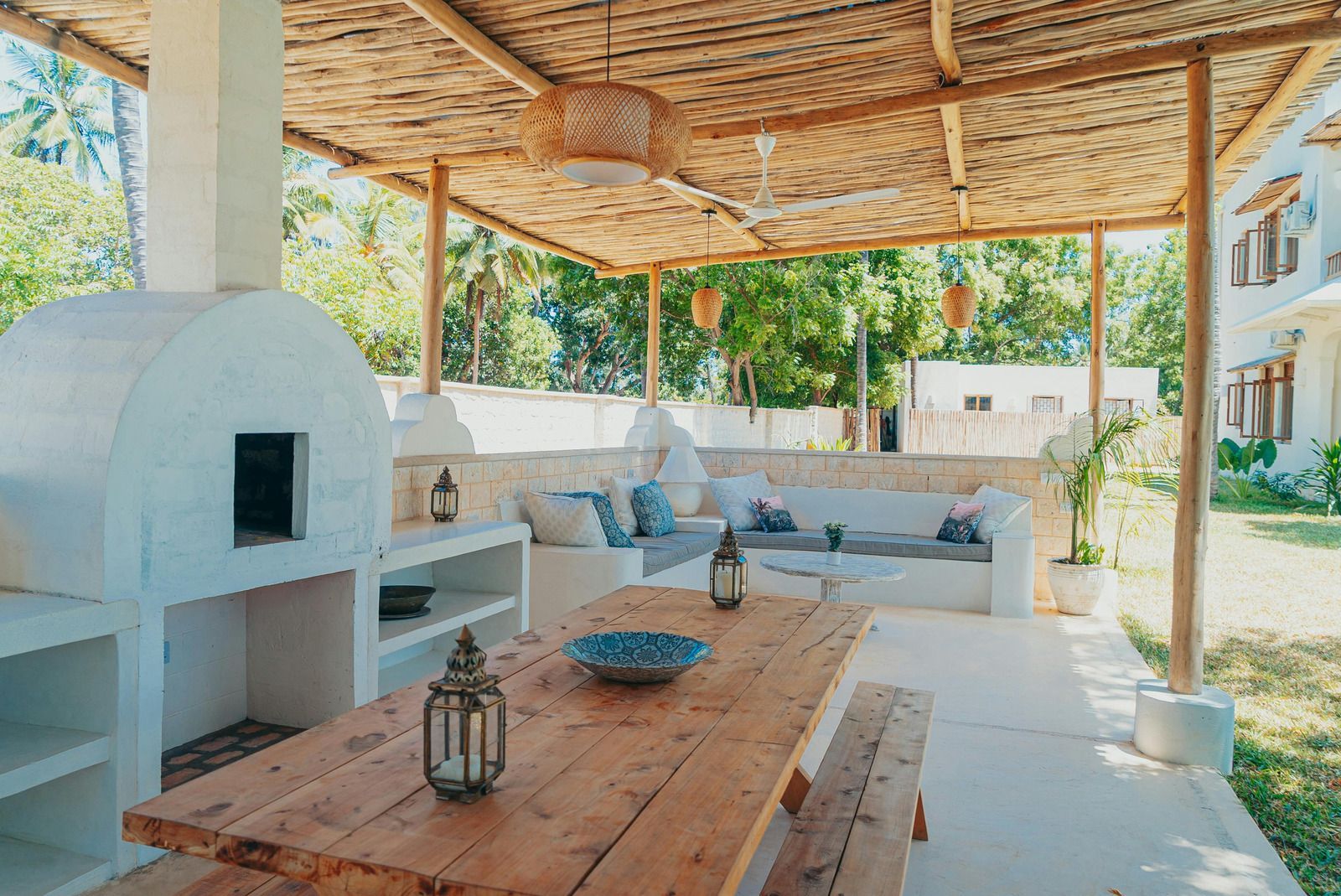 Outdoor dining area with a wood table, built-in seating, and pizza oven, under a thatched roof.