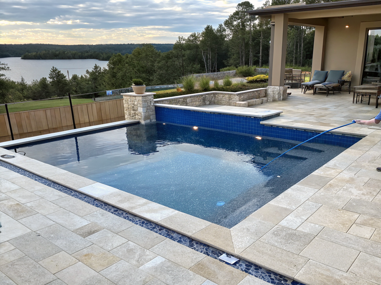 Rectangular blue tile pool with stone patio, overlooking a lake and trees.
