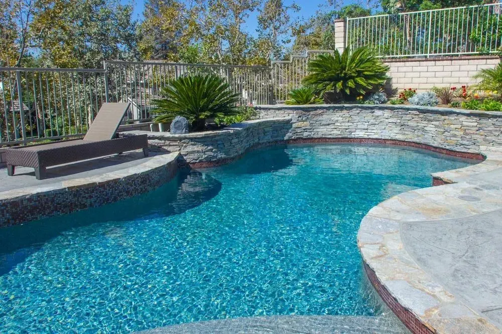 Swimming pool with turquoise water and stone border, surrounded by landscaping and a lounge chair.