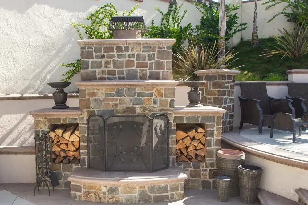 Stone outdoor fireplace with stacked firewood, patio, and chairs.