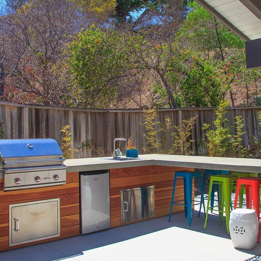 Outdoor kitchen with grill, sink, refrigerator, and colorful bar stools. Wooden paneling.