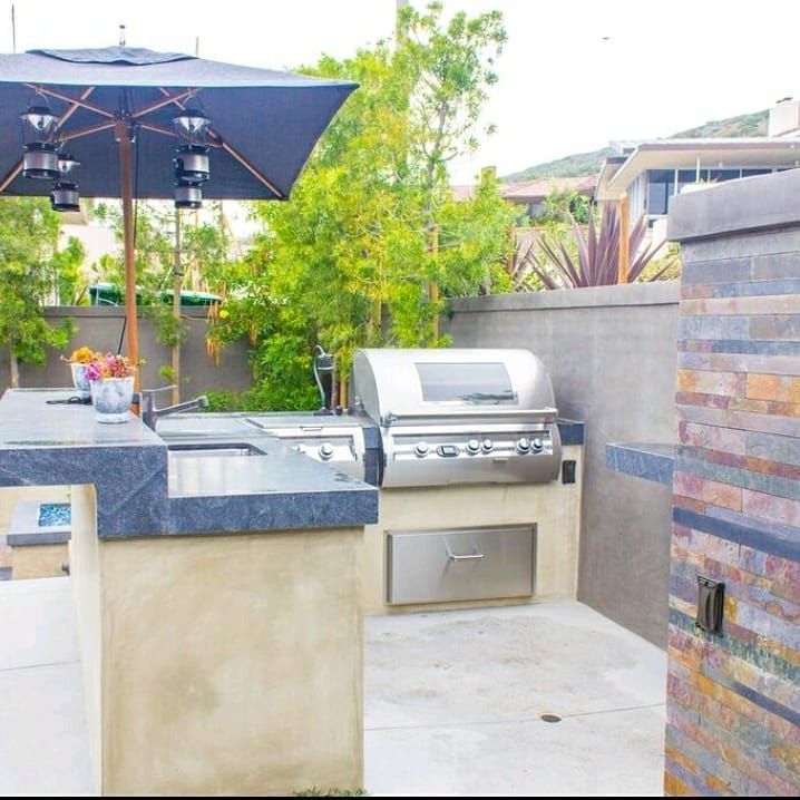 Outdoor kitchen with grill, sink, and countertop under umbrella. Beige and gray color scheme.