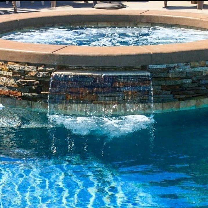A swimming pool with a hot tub and waterfall feature, blue water, stone accents.