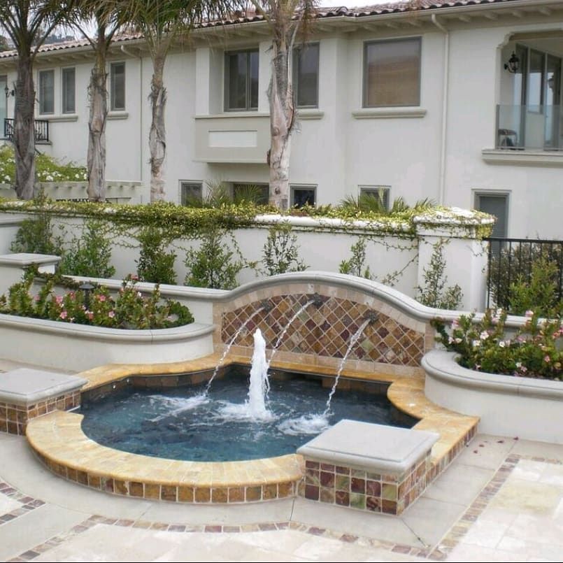 Water fountain in a courtyard with tiered planters and a light-colored building in the background.
