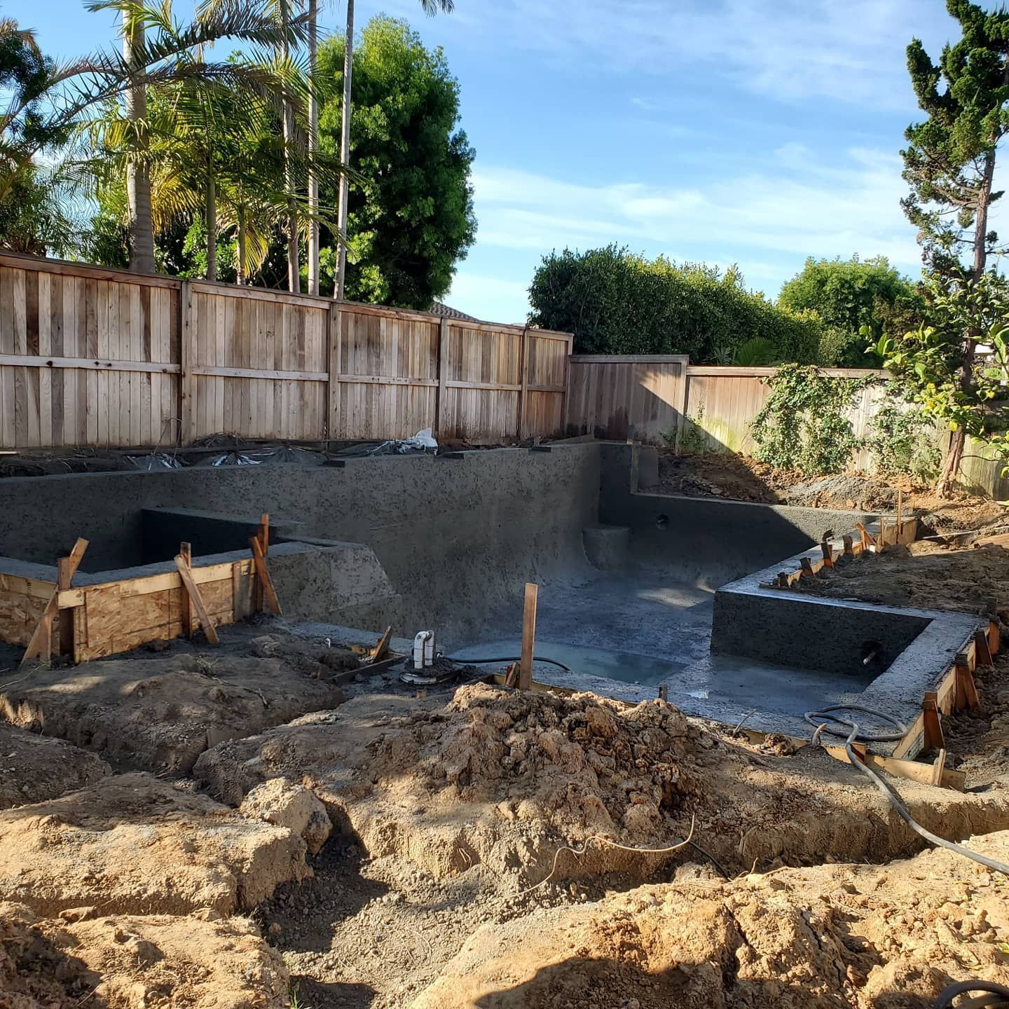 Pool construction site with concrete forms, soil, and a wooden fence.