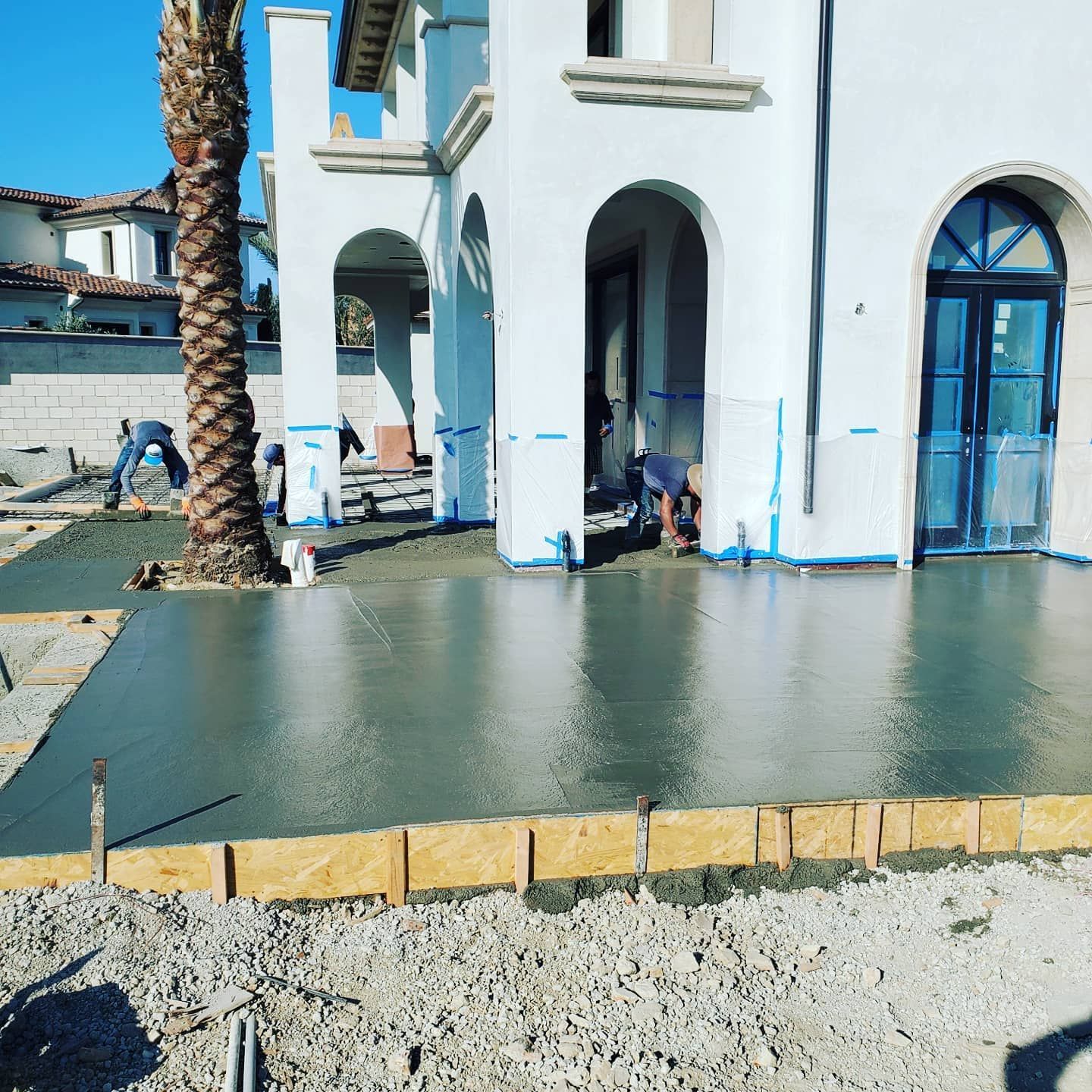 Workers pouring concrete patio in front of a white house with arches and a palm tree.