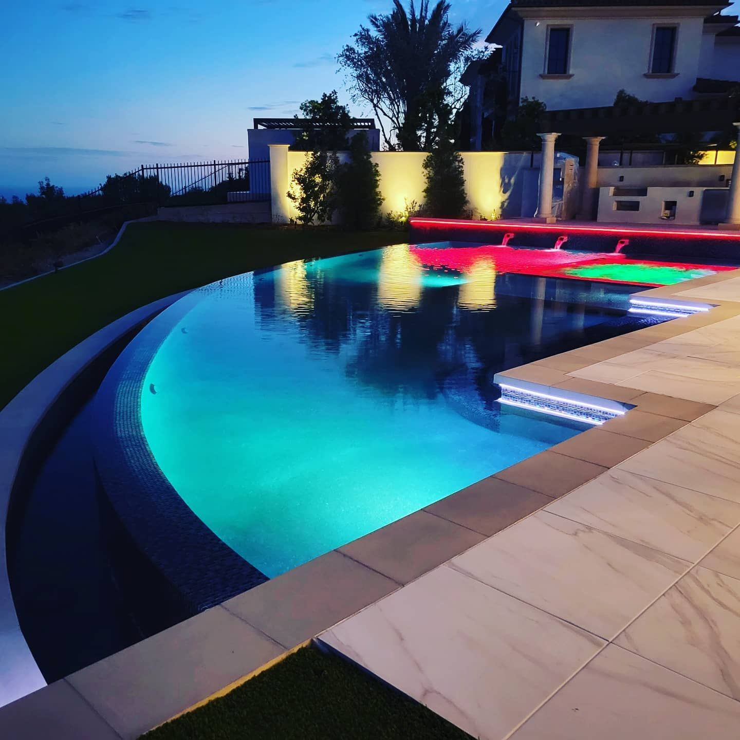 Illuminated infinity pool with colorful lights at dusk. Modern home in background.