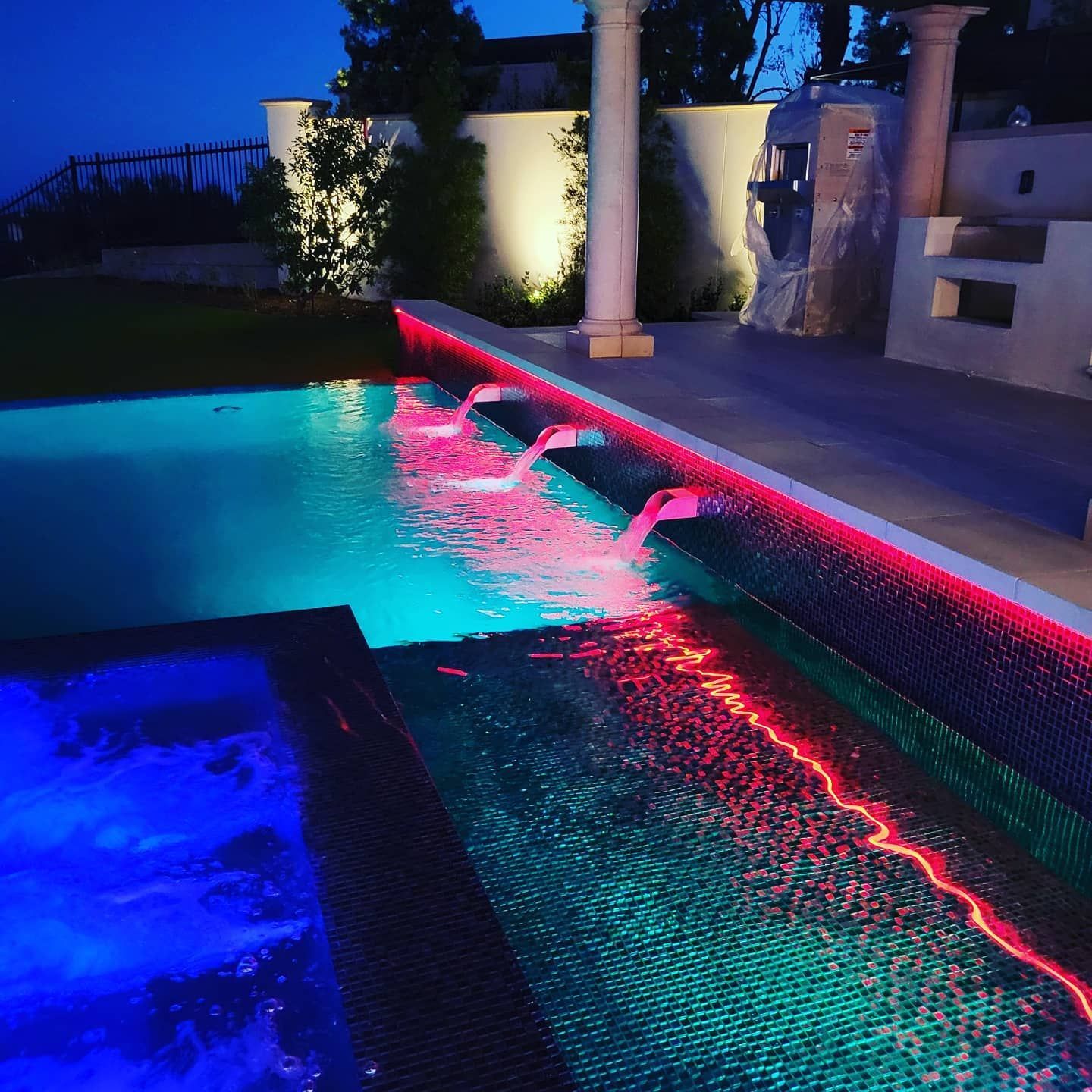 Illuminated swimming pool at night with cascading water and colored lights.