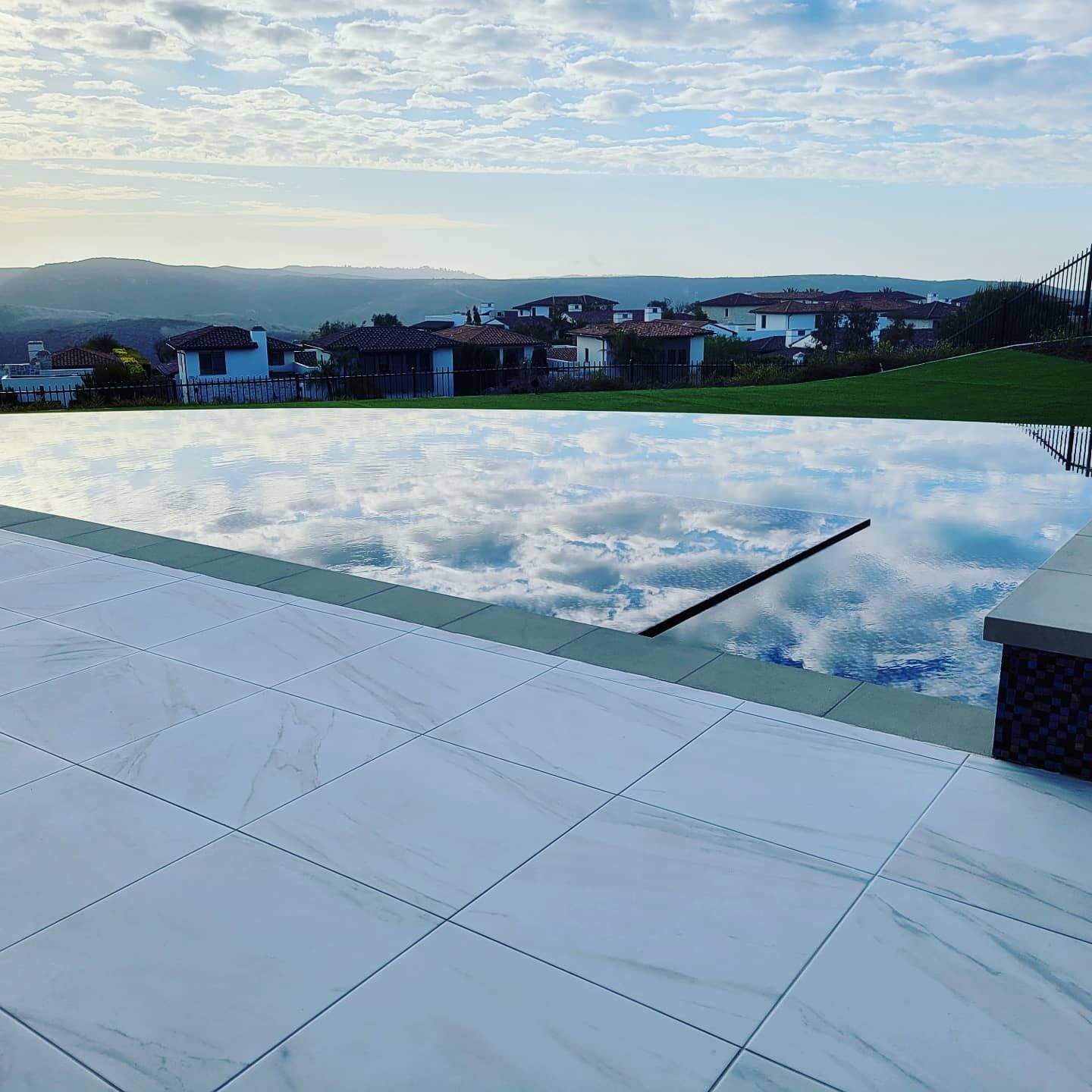 An infinity pool reflects a cloudy sky over a residential area, with white tiled patio in foreground.