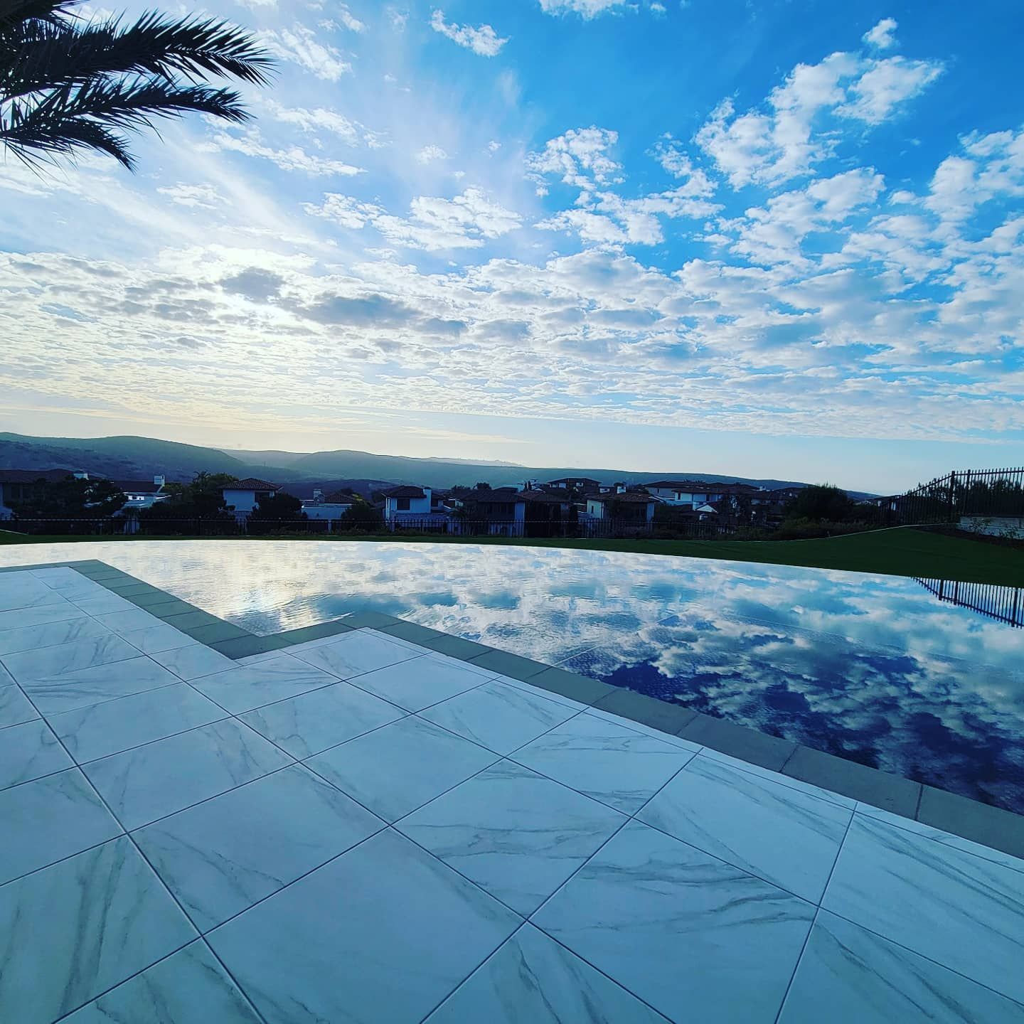 A swimming pool reflects the cloudy sky and a distant town under a bright blue sky.