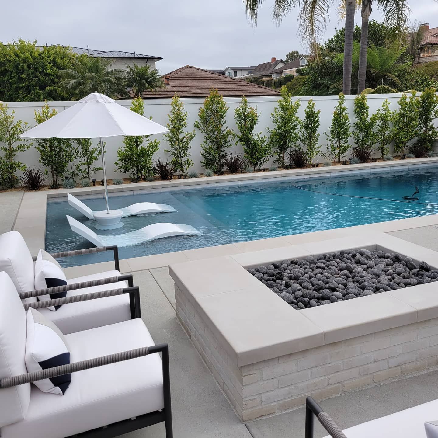 Poolside patio with white lounge chairs, fire pit, and umbrella. Pool in the background with two floating chaise lounges.