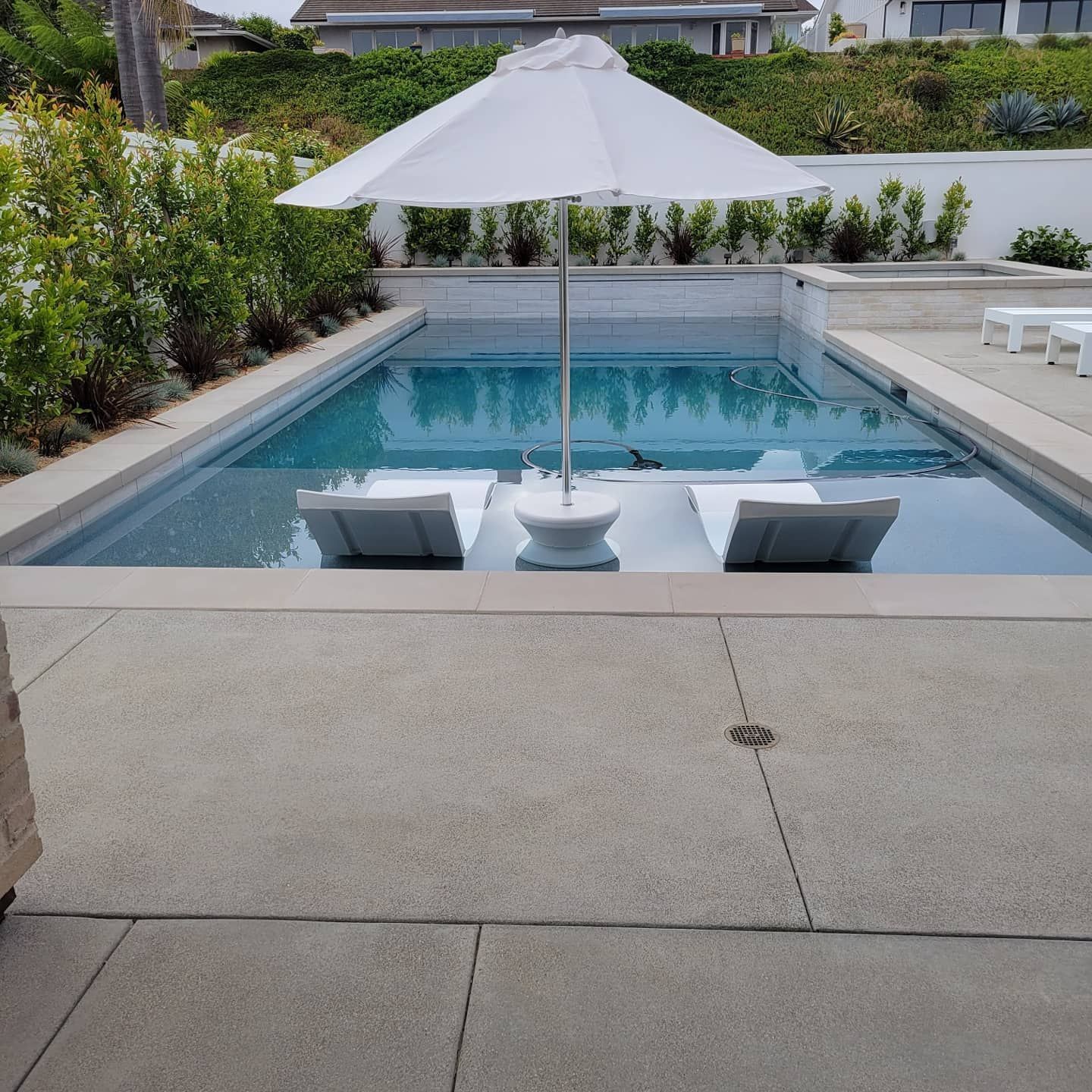 Pool with lounge chairs and umbrella. Stone patio. Green shrubs and white building in the background.