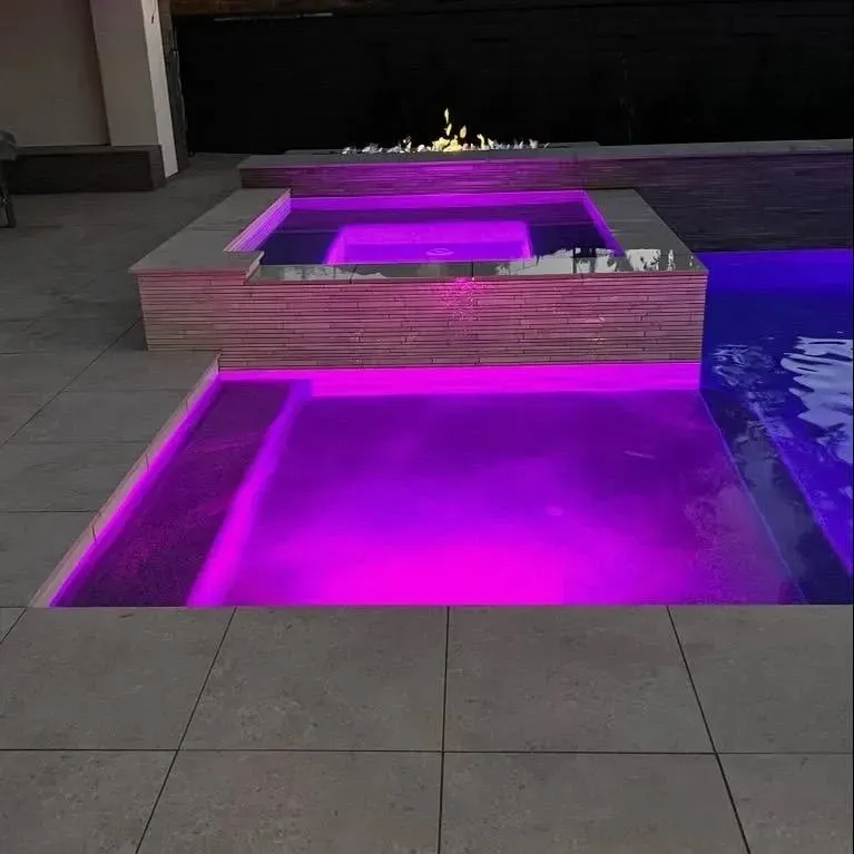 A pool and spa with purple LED lights, outdoor setting. Fire pit in the background.
