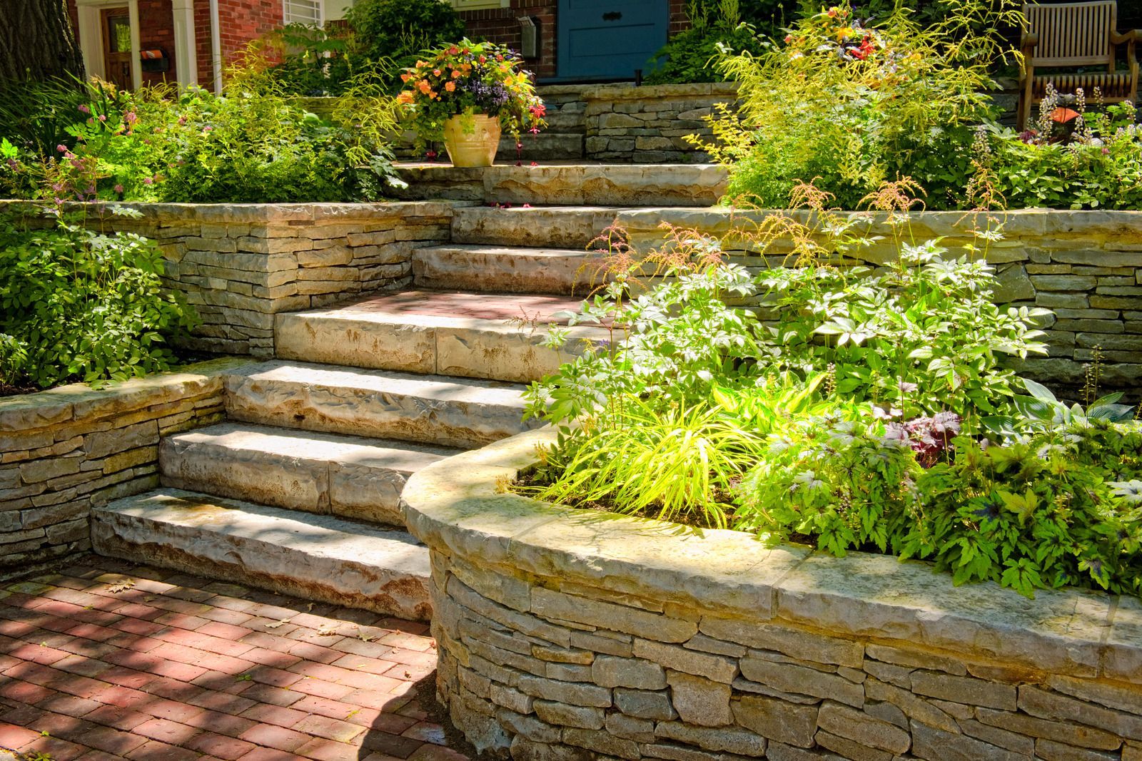 Stone steps ascend, surrounded by lush green plants and flowers. Red brick patio.
