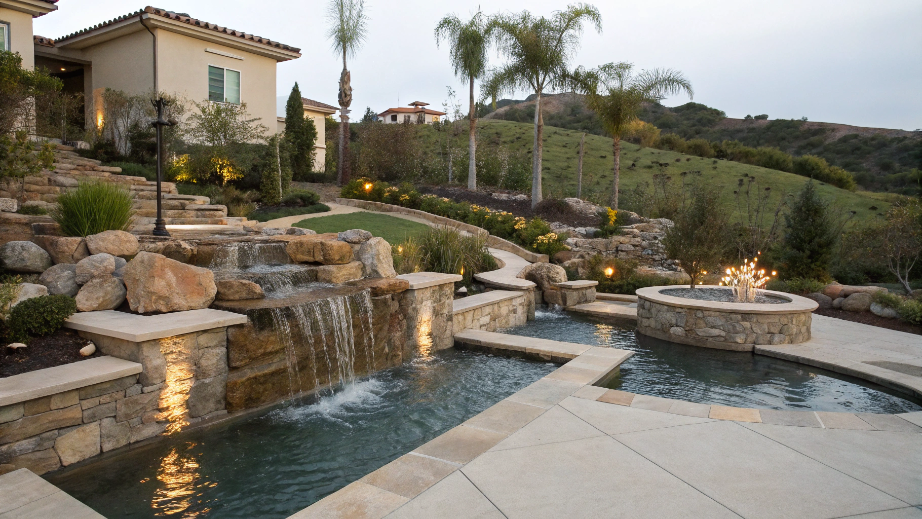 Waterfalls cascade into a pool in a luxury backyard. Stone features, greenery, and hill in the background.