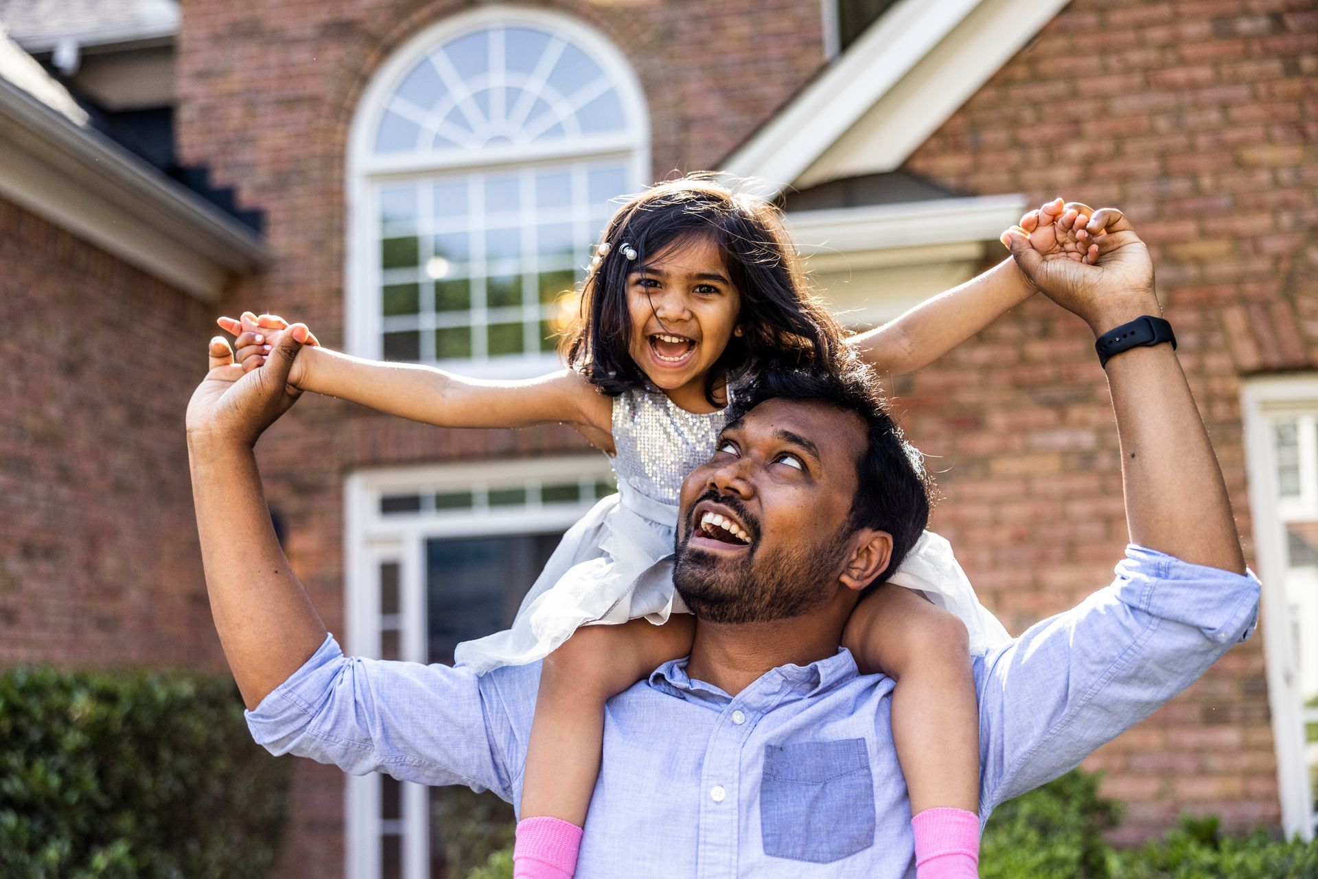 A man is carrying a little girl on his shoulders in front of a brick house.