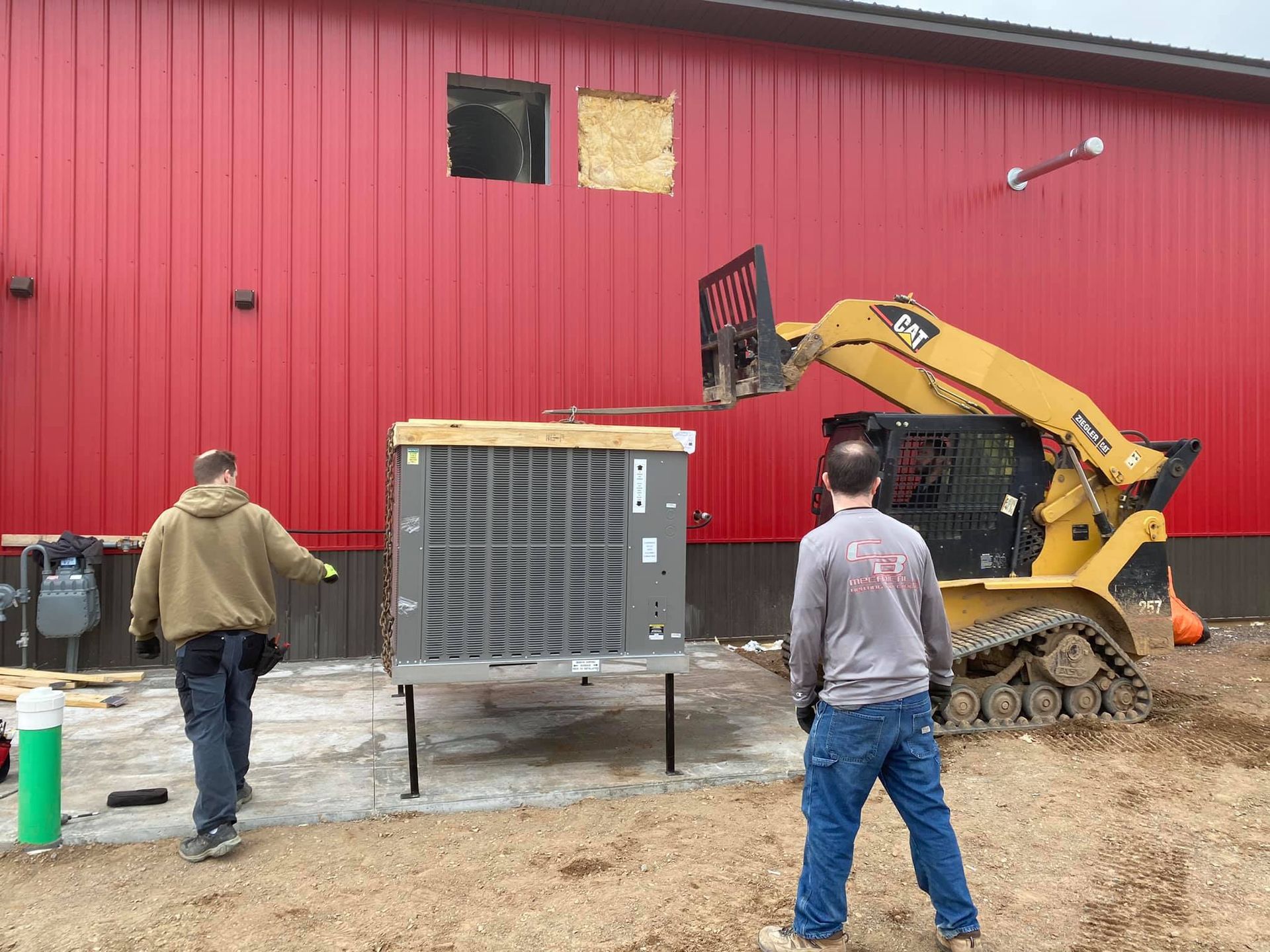 Workers installing HVAC unit near red building with skid steer.