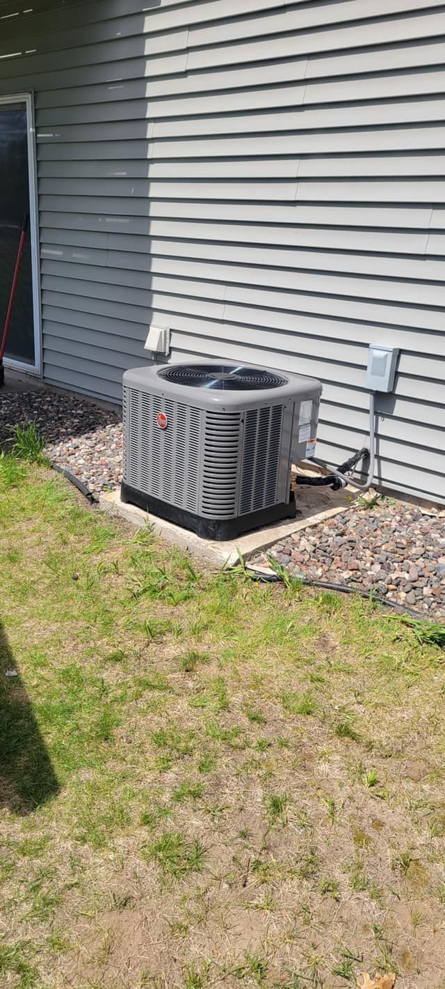 An air conditioning unit next to a gray-sided building sits on a gravel bed and green lawn.