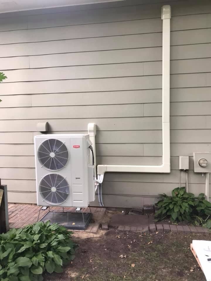 Exterior heat pump unit mounted on a light-colored building with white ducting and landscaping.