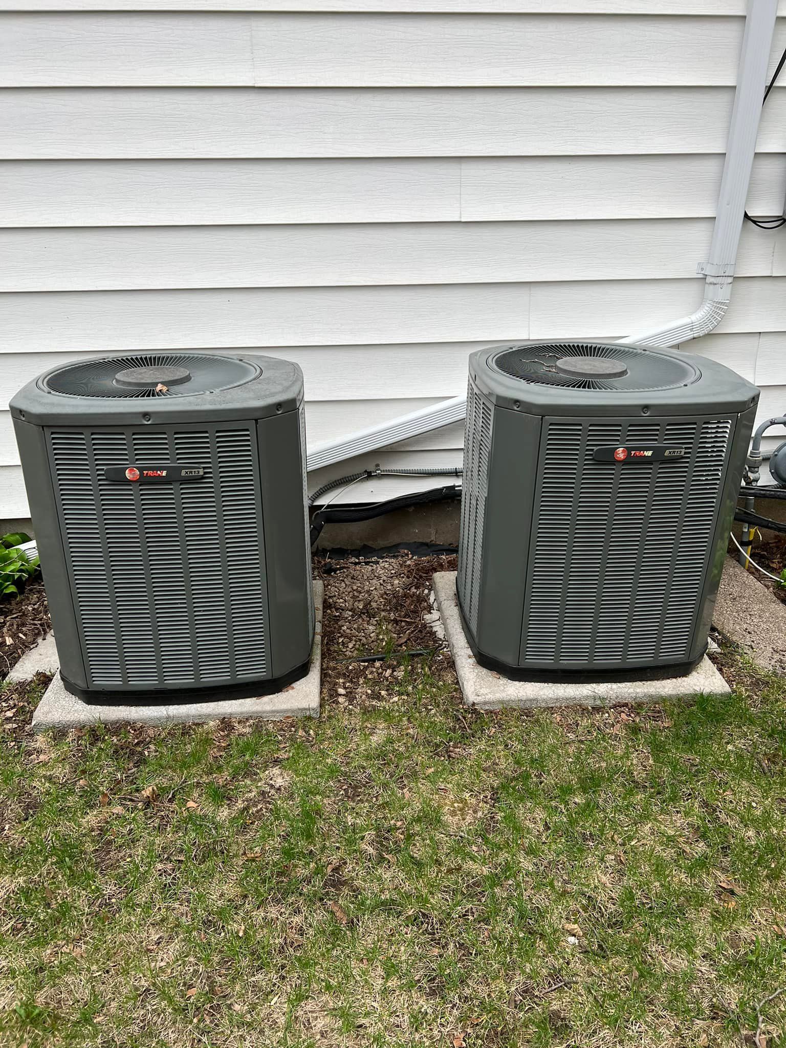 Two gray air conditioning units sitting on concrete pads next to a white house.