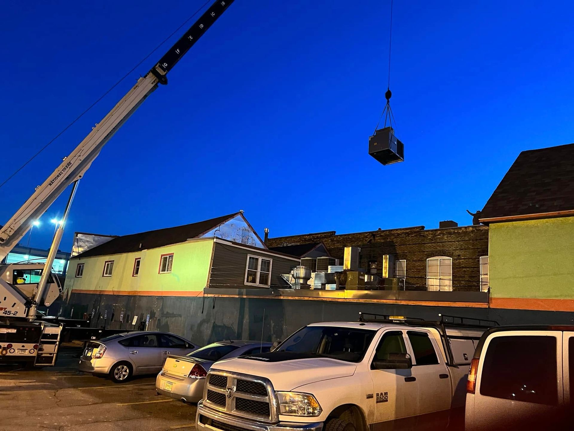 A crane lifting a dark object above a building with a blue sky background. Cars are parked in the foreground.