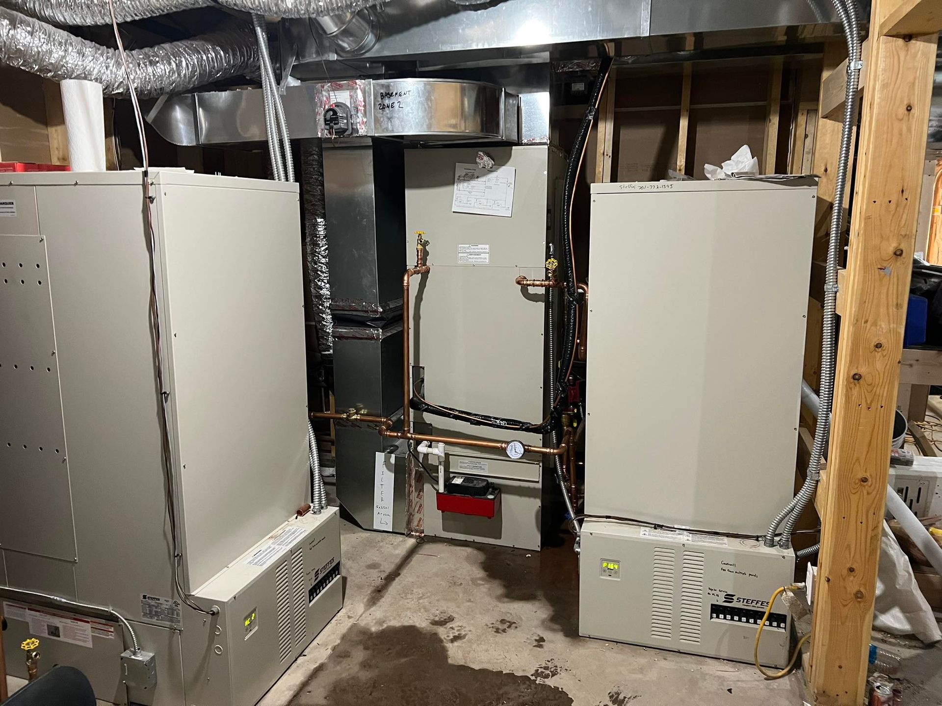 Two HVAC units and related piping installed in a basement, with exposed wooden framing and ductwork.