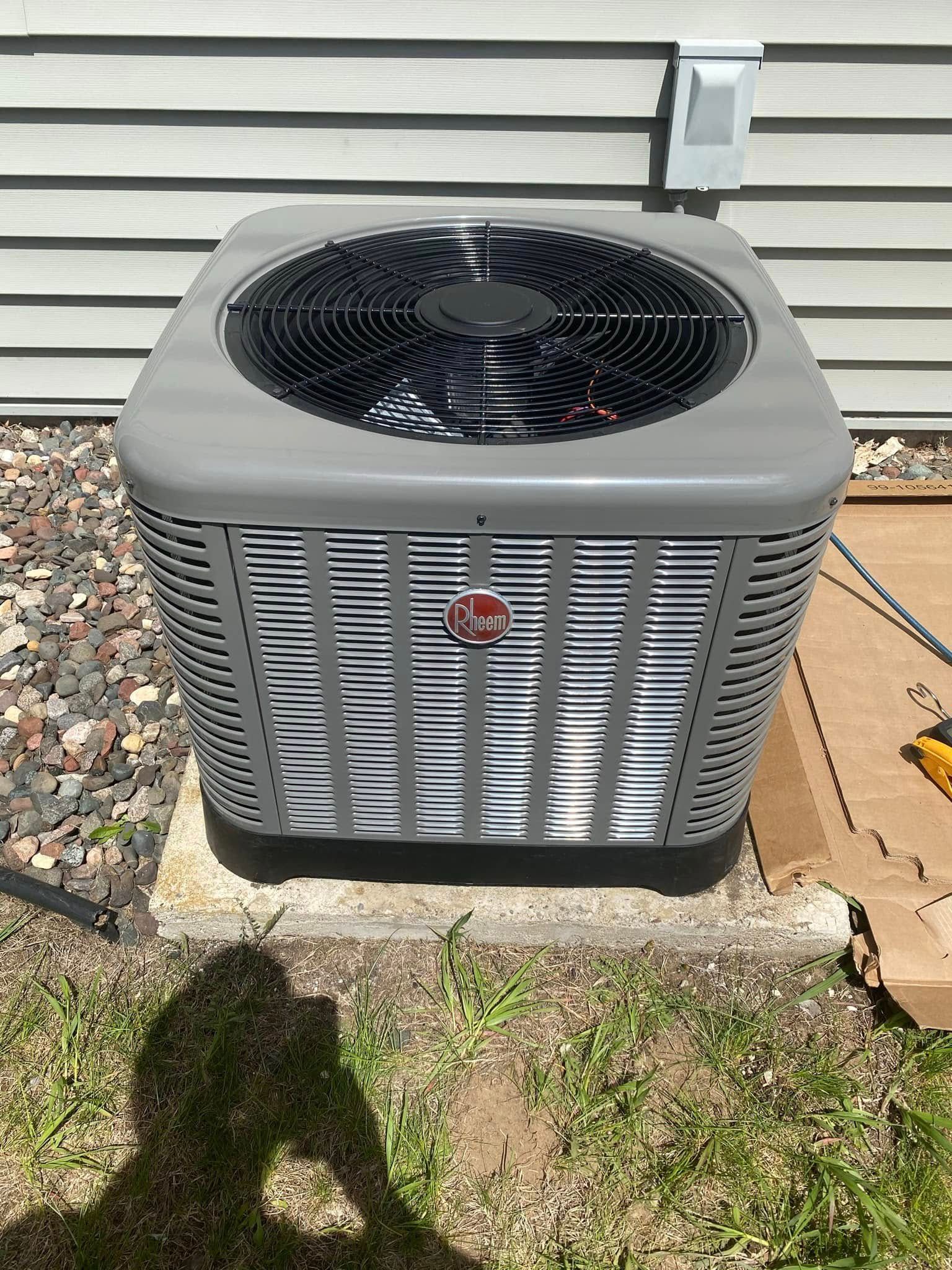 Gray Rheem air conditioning unit on a concrete pad near a house, with a shadow of a person.