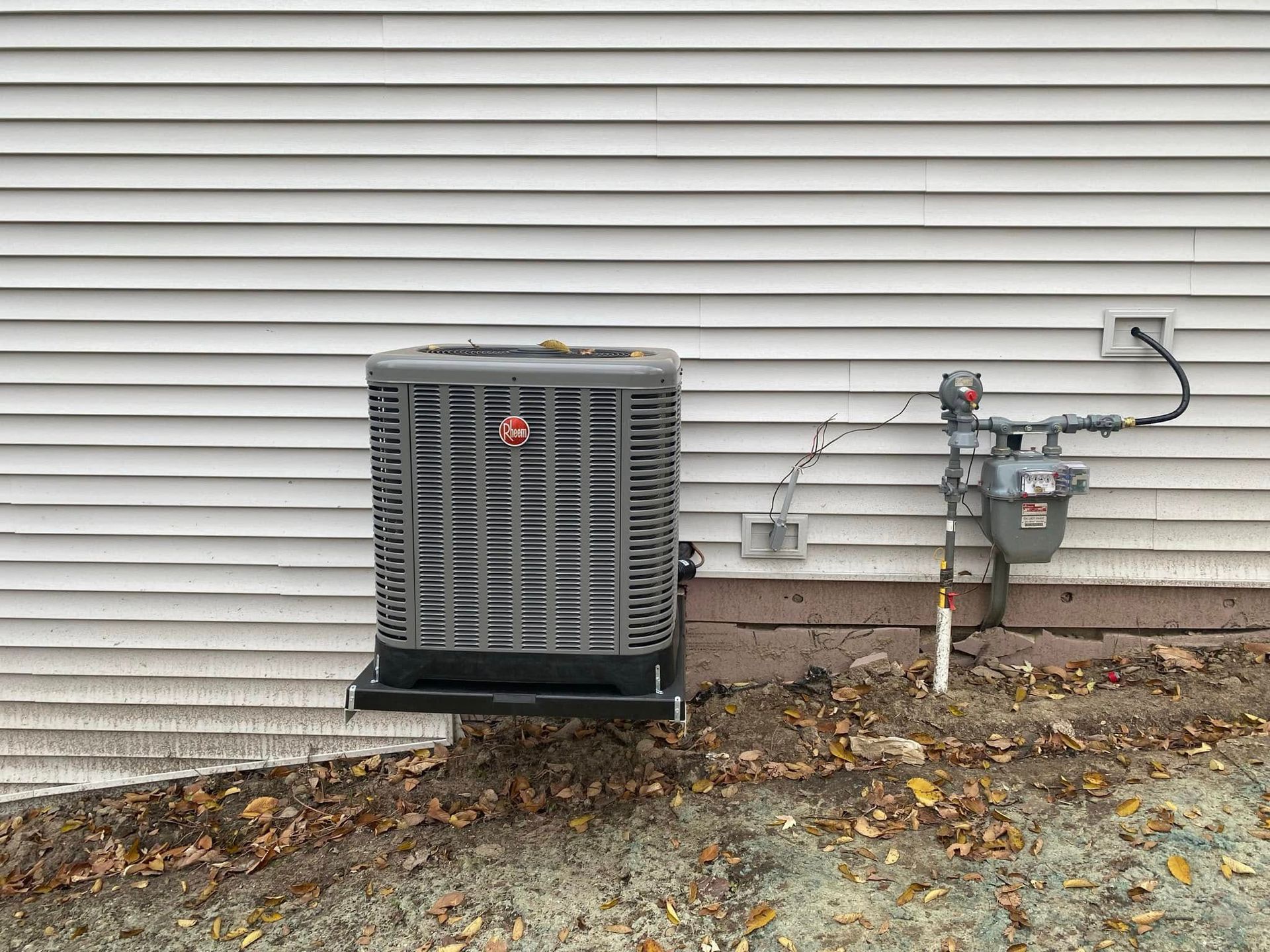 Air conditioning unit outside a white-sided building next to a gas meter, set in autumn.