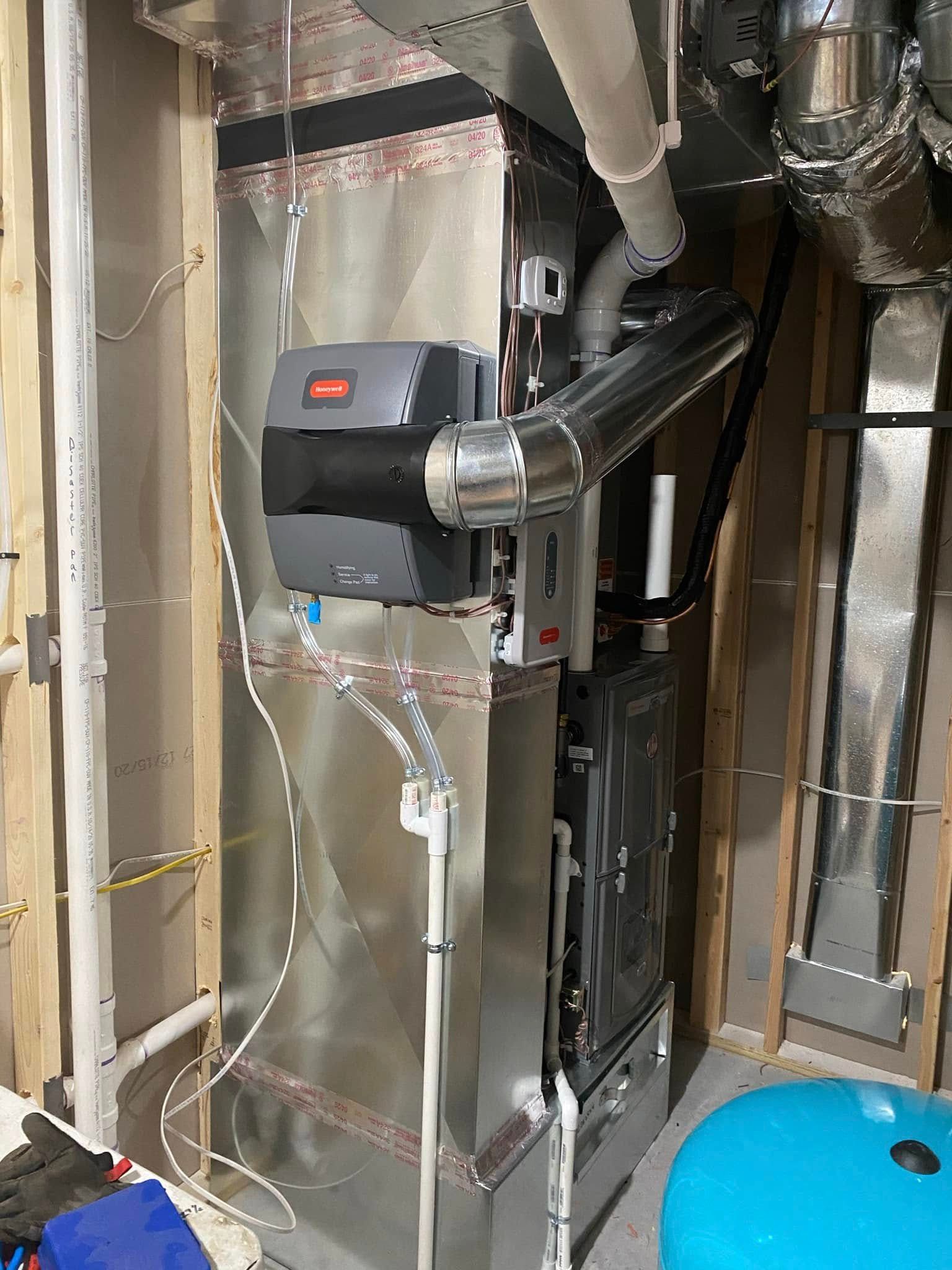 A humidifier unit attached to a furnace in a basement setting. Pipes and wiring visible.