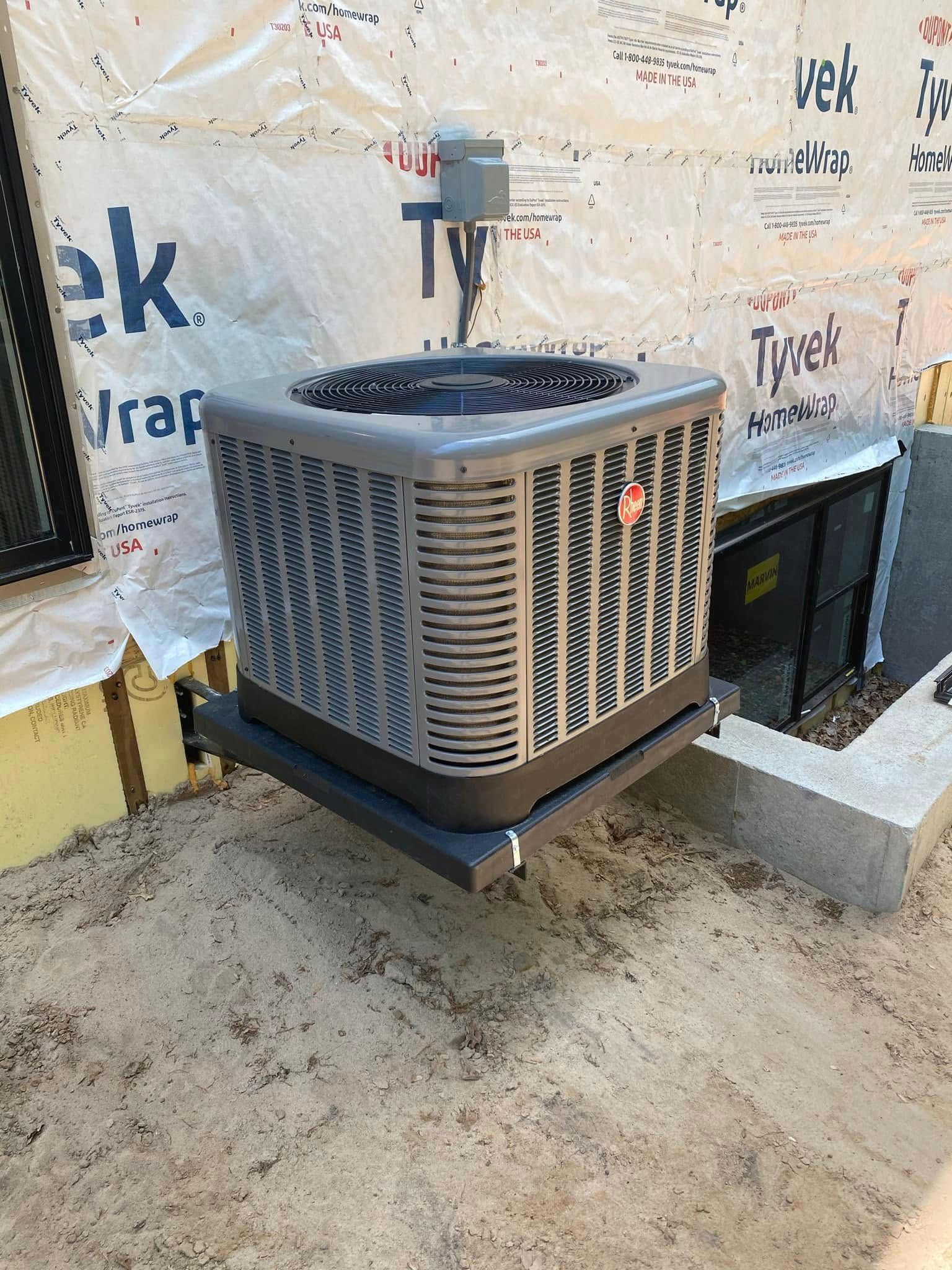 Rheem air conditioning unit mounted on a black platform against a building, next to a concrete block.