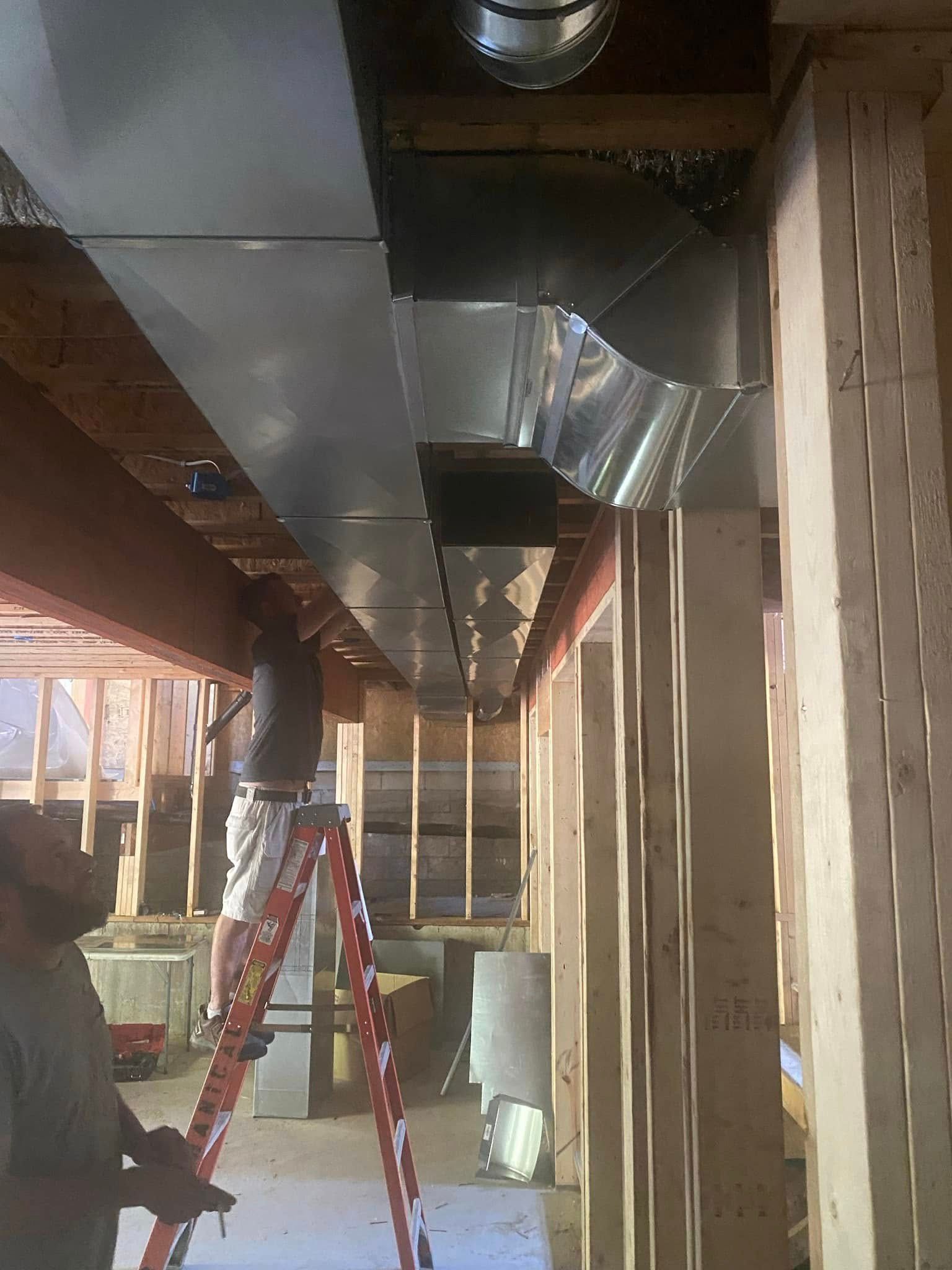 Two workers installing HVAC ductwork in a basement; one on a ladder.