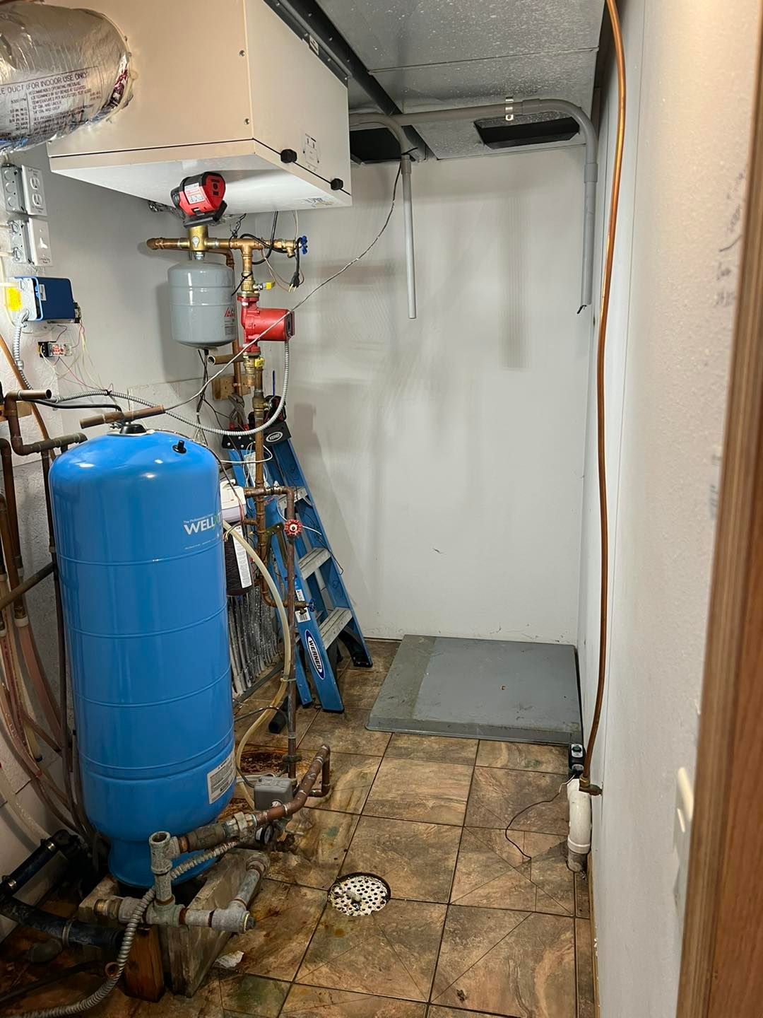 Basement mechanical room with blue water tank, pipes, and a ladder.