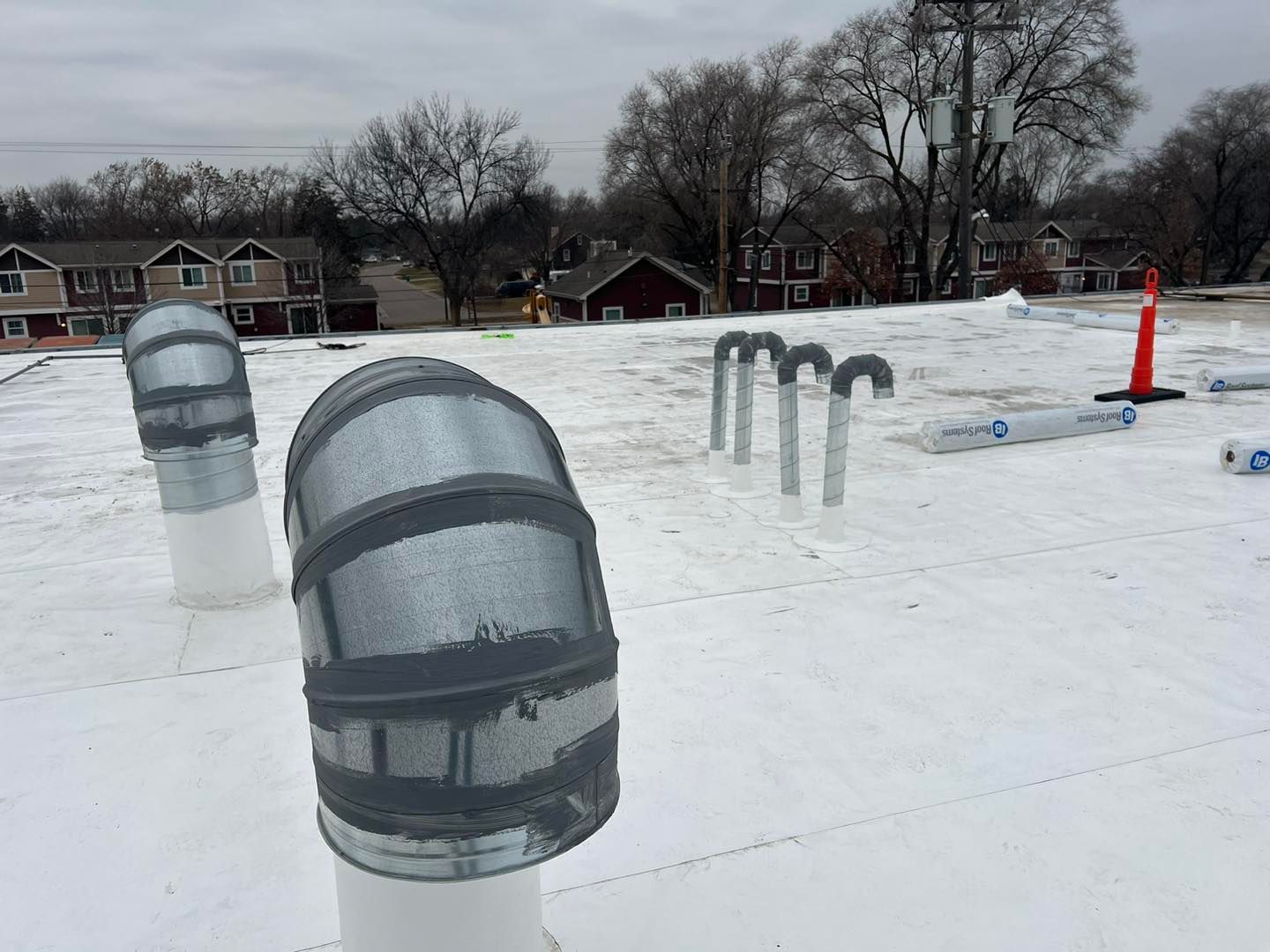 White flat roof with metal vents and pipes, overcast sky, residential buildings in background.