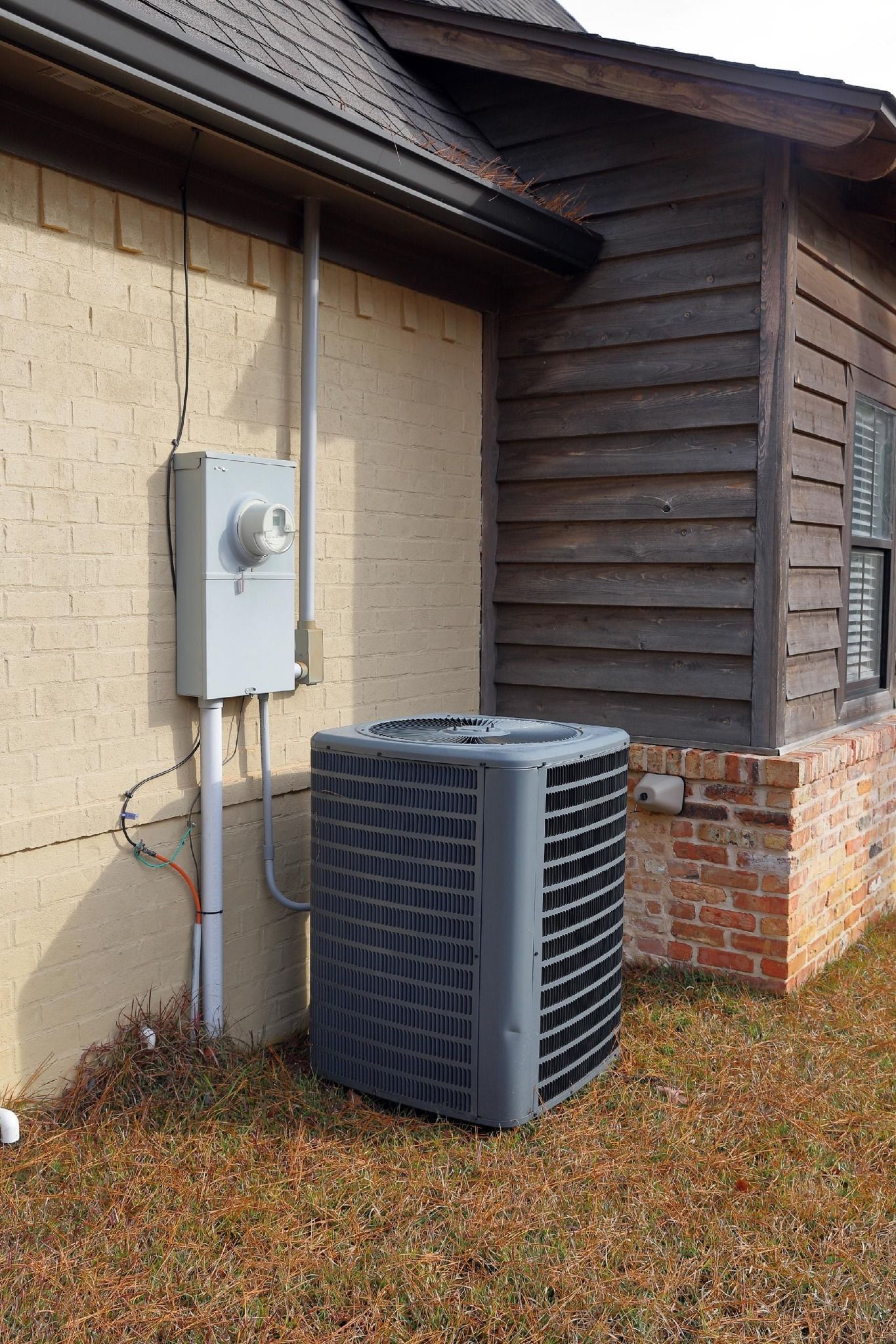 Air conditioning unit next to a brick and wood-sided building, with electrical box.