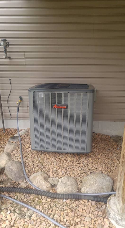 A gray Trane air conditioning unit sits on gravel near a building. A hose and rocks surround it.