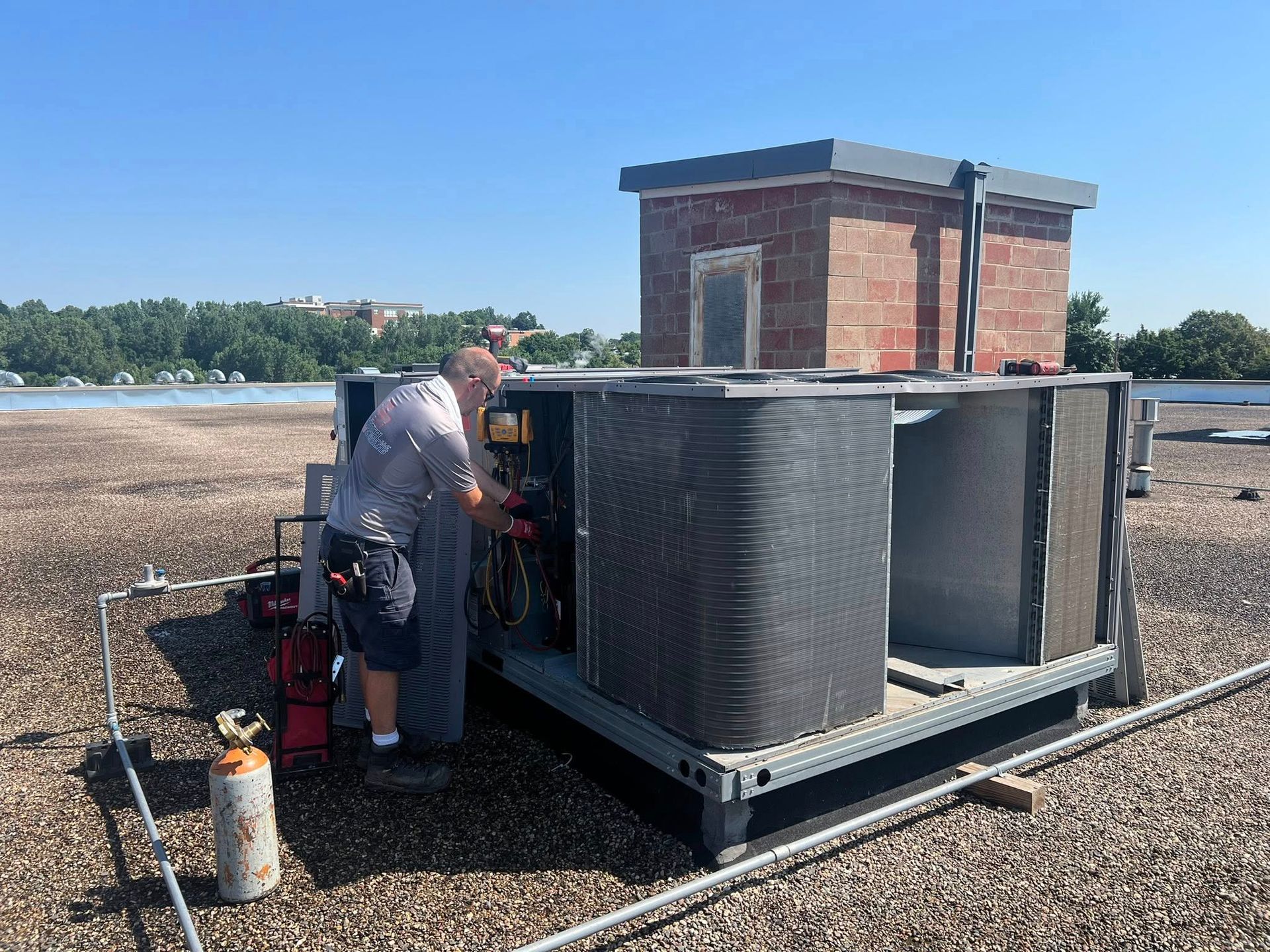 HVAC technician working on rooftop air conditioning unit, next to small brick structure under a bright blue sky.