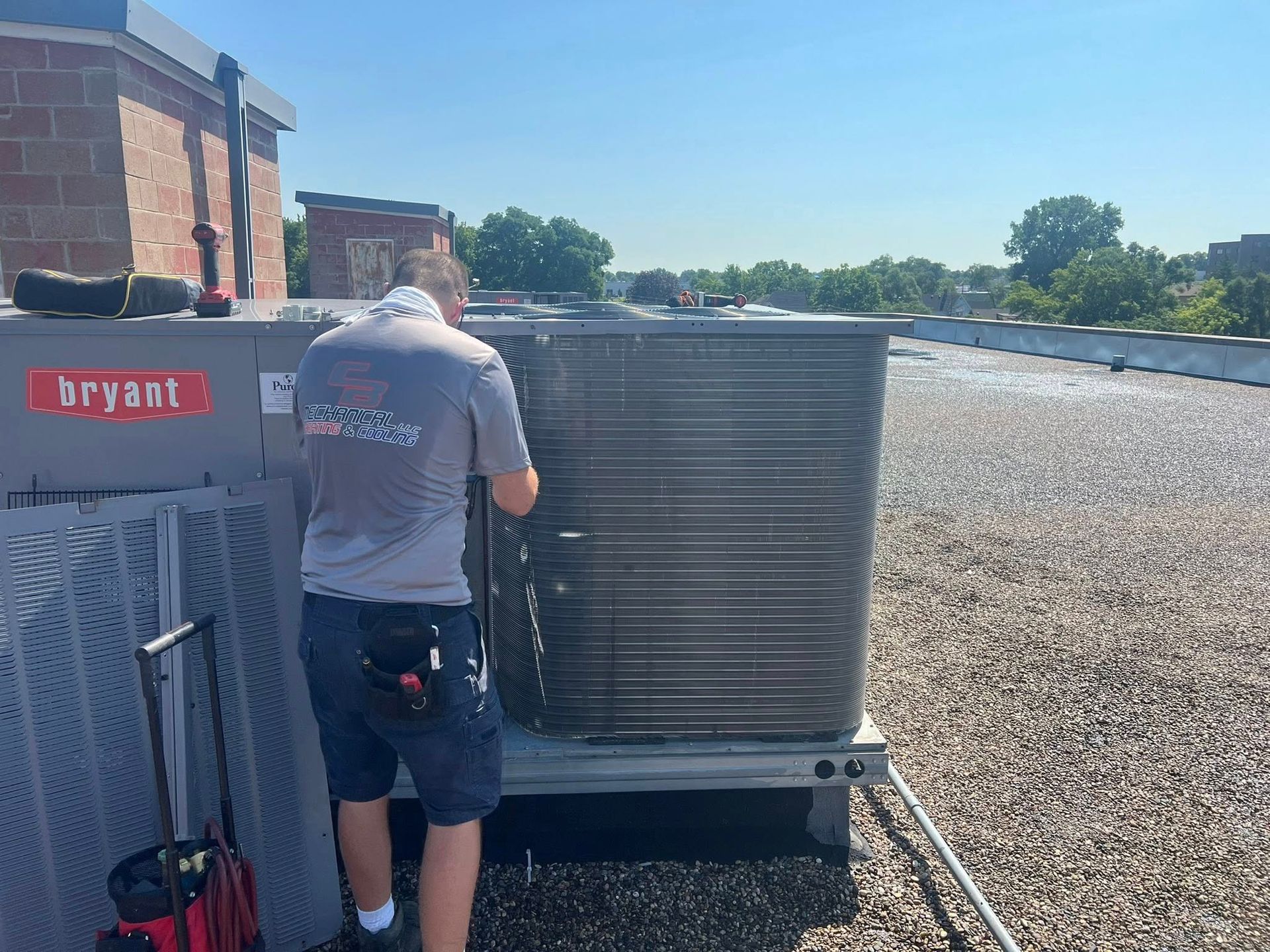 A technician cleans an air conditioning unit on a rooftop under a blue sky.