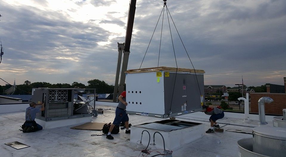 A crane lifting a large HVAC unit onto a white rooftop. Workers are guiding it. Cloudy sky.