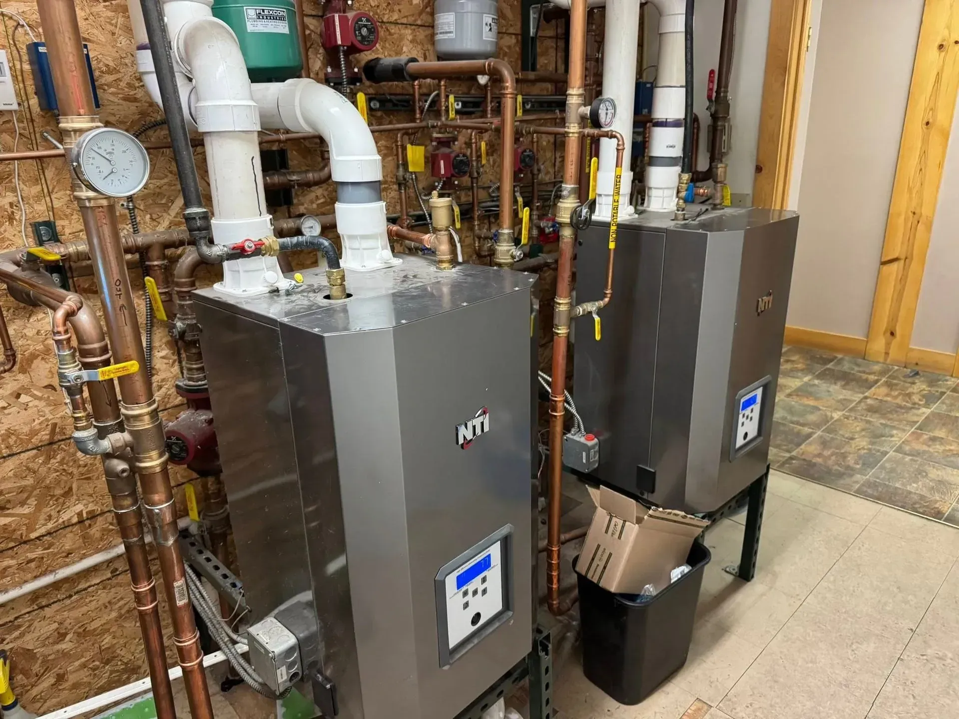 Two silver wall-mounted heating units with white pipes and copper plumbing in a utility room.